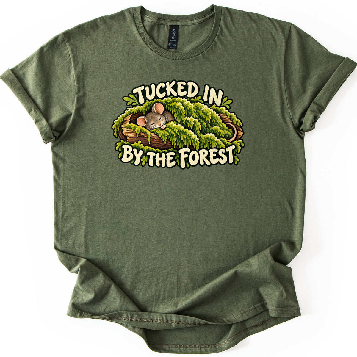 Tucked in by the Forest Comfort Colors T-shirt in Military Green, laid flat. A sleeping gray mouse with pink ears lies under a blanket of green moss in a hollow log, between cream words reading Tucked in By the Forest. The shirt has rolled sleeves on a white background.