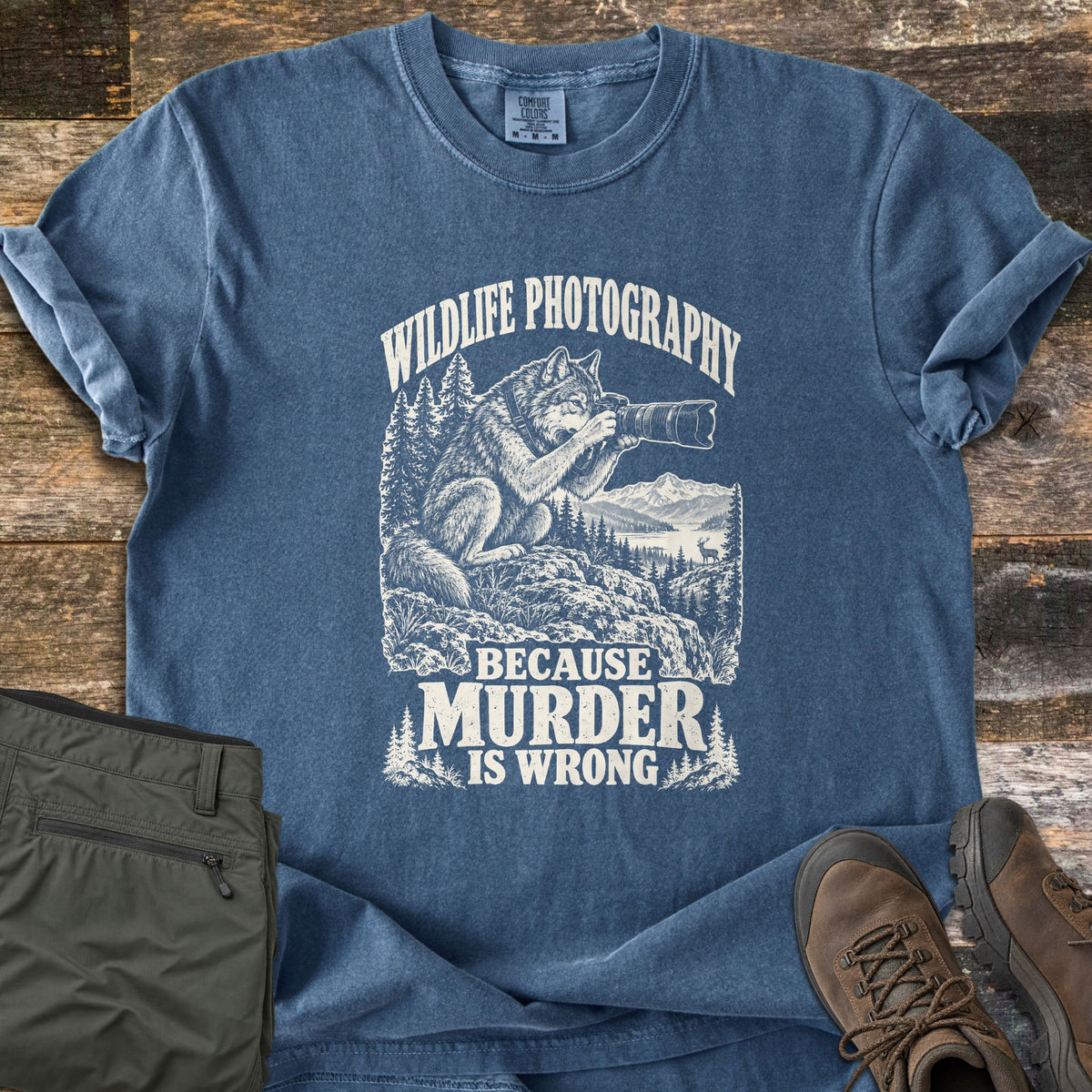 Wildlife Photography Because Murder Is Wrong Comfort Colors T-shirt in Denim, laid flat. White ink shows a wolf on a rocky ledge aiming a long-lens camera at a distant deer by a mountain lake, under Wildlife Photography and above Because Murder Is Wrong. Olive pants and brown hiking boots lie on dark wood.