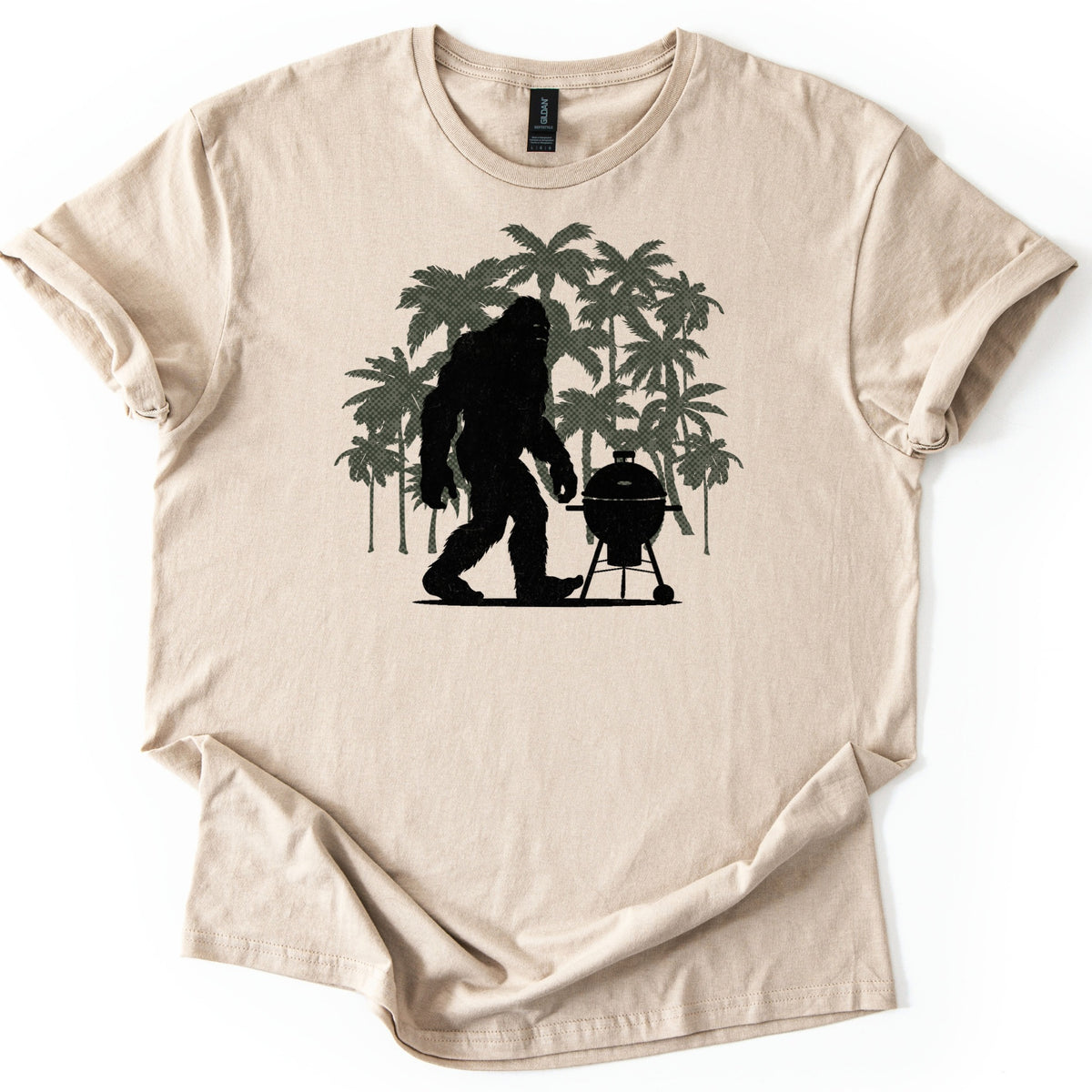 Squatch BBQ Smoker Comfort Colors T-shirt in Sand, laid flat. A black silhouette of Bigfoot walks beside a kettle-style grill on wheeled legs, set against a cluster of olive green palm trees. No words are printed. The shirt lies on a white surface with sleeves cuffed.