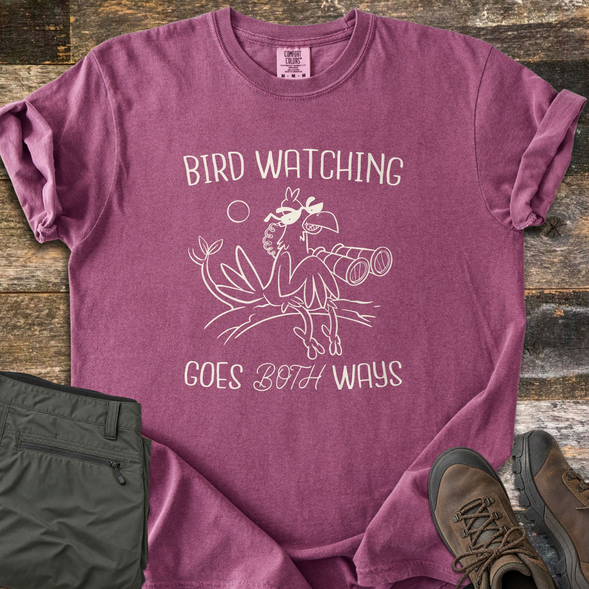 Birds Watch Back Birder Comfort Colors T-shirt in Berry, laid flat. A cartoon bird in sunglasses perches on a branch beside binoculars between the words Bird Watching Goes Both Ways in cream. Olive hiking pants and brown hiking boots sit beside it on weathered wood.