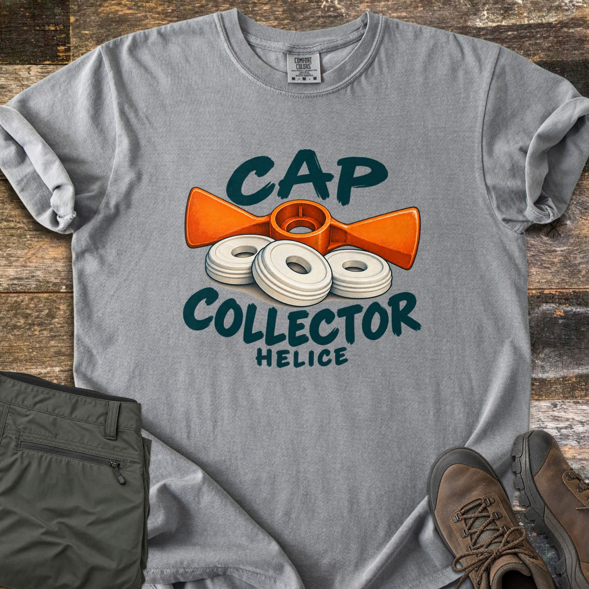 Cap Collector Comfort Colors T-shirt in Grey, laid flat. An orange helice key and three white caps sit between the words Cap Collector, with Helice below. Sleeves are rolled; olive pants and brown hiking boots lie beside it on dark wood planks.