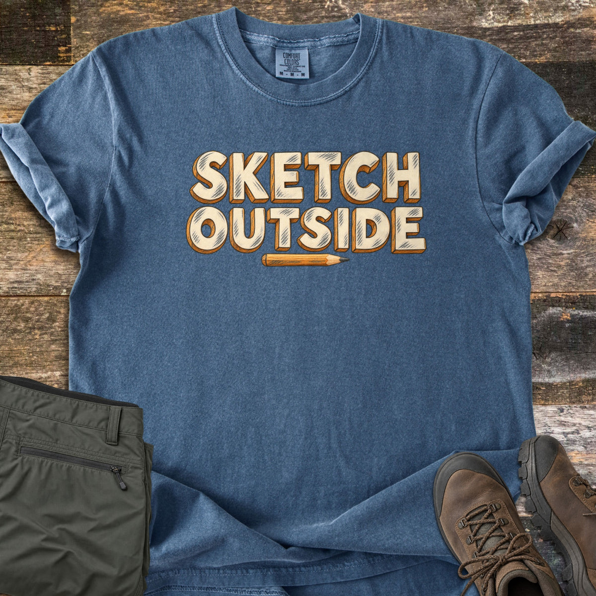 Sketch Outside Comfort Colors T-shirt in Denim, laid flat. Cream block letters with pencil-style shading read Sketch Outside above a drawn yellow pencil. Sleeves rolled on a weathered wood floor beside folded olive hiking pants and brown leather hiking boots.