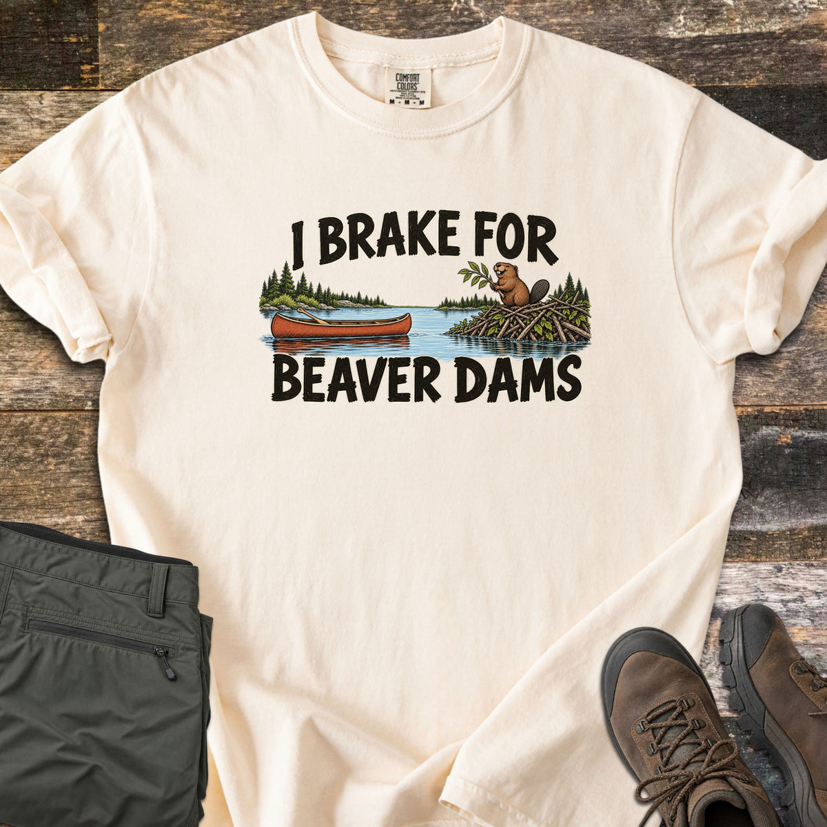 Brake For Beaver Dams Comfort Colors T-shirt in Ivory, laid flat. The words I Brake For Beaver Dams frame a lake scene with a red canoe and a beaver on its stick dam among pine trees. Sleeves rolled, on weathered wood with folded green hiking pants and brown hiking boots.