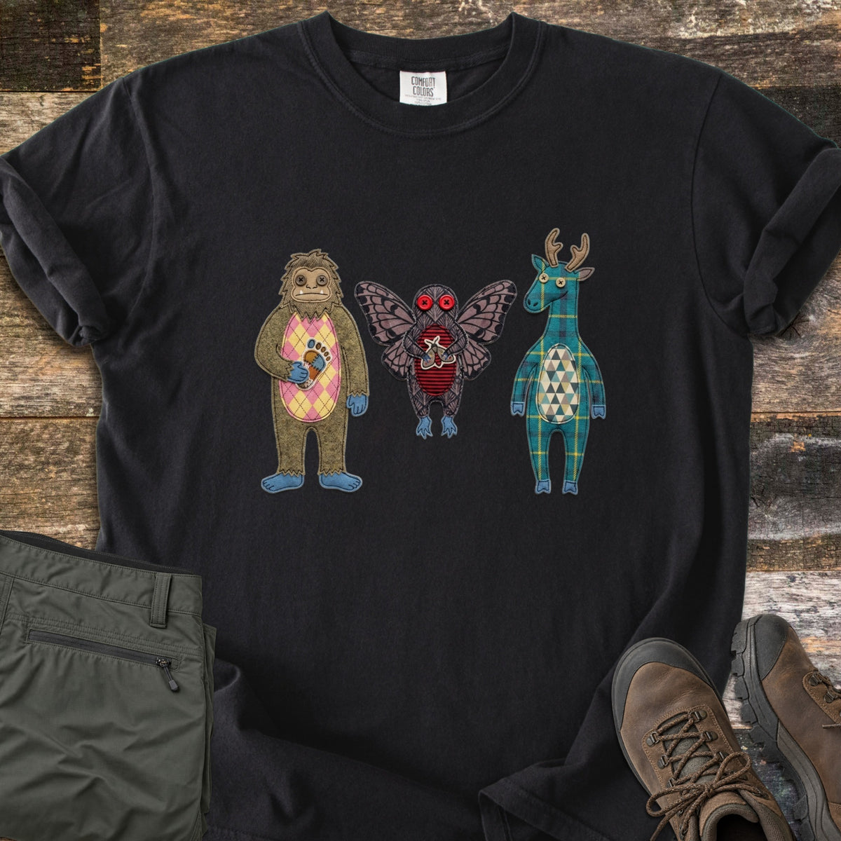 Patchwork Cryptid Trio Comfort Colors T-shirt in Black, laid flat. Three patchwork fabric cryptids: Bigfoot with a pink argyle belly holding a footprint patch, a red-eyed Mothman, and a plaid jackalope. Laid on weathered wood beside olive pants and brown hiking boots.