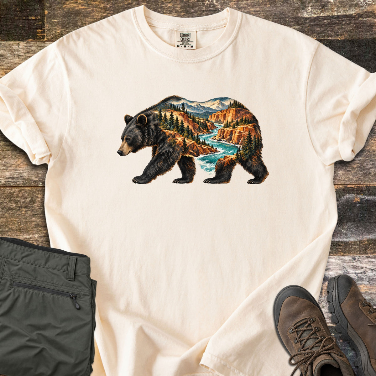 Bear River Canyon Comfort Colors T-shirt in Ivory, laid flat. A walking black bear whose body is filled with a canyon scene of orange cliffs, pine trees, a turquoise river and snowy peaks. Folded olive hiking pants and brown hiking boots sit on weathered wood boards.