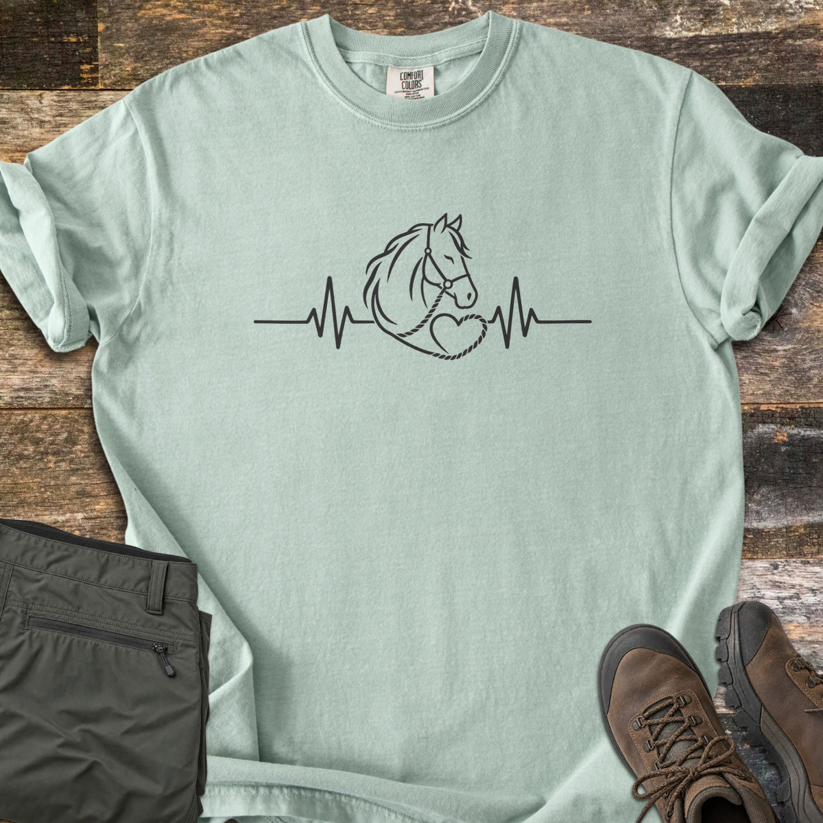 Horse Heartbeat Comfort Colors T-shirt in Bay, laid flat. A black line drawing of a horse head in a halter, its lead rope looping into a heart, sits on a heartbeat line. Sleeves rolled, on weathered wood with folded olive pants and brown hiking boots.