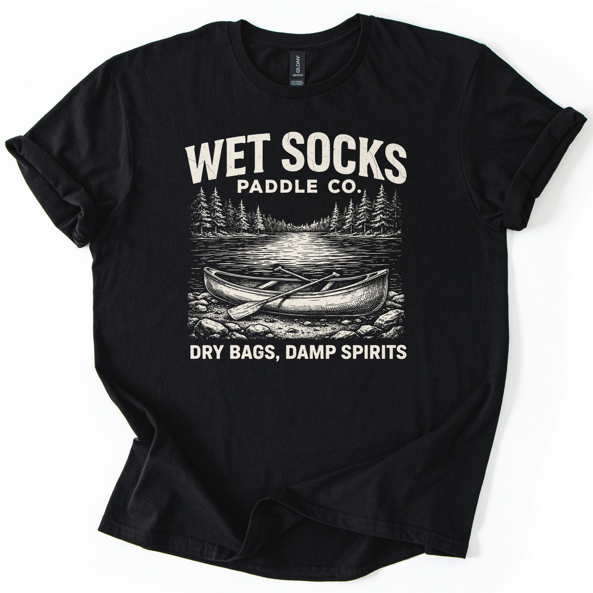 Wet Socks Paddle Co Comfort Colors T-shirt in Black, laid flat. A cream etched drawing of a canoe with crossed paddles on a rocky shore, with a lake and pine trees behind, under the words Wet Socks Paddle Co. and above Dry Bags, Damp Spirits. White background.