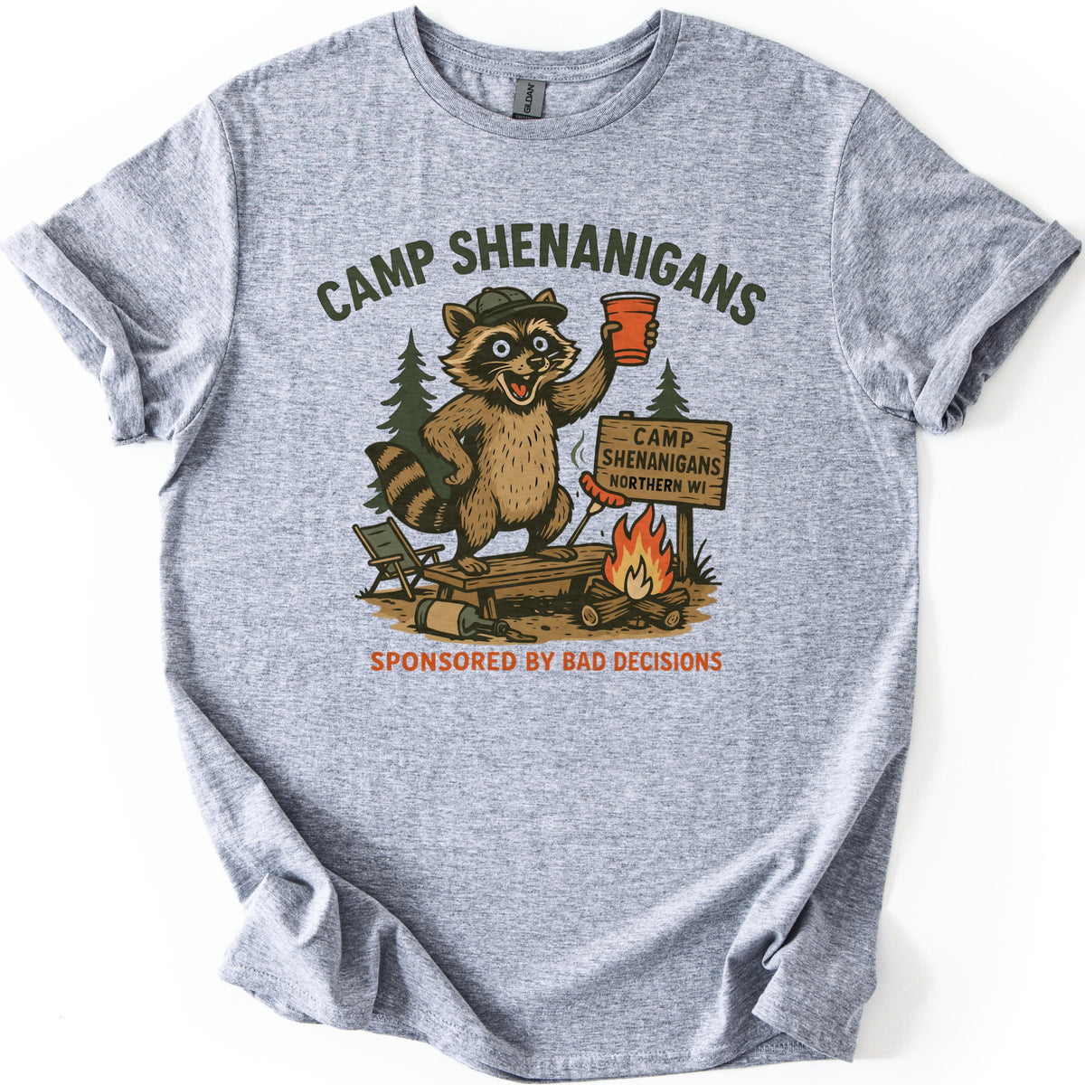 Camp Shenanigans Comfort Colors T-shirt in Sport Grey, laid flat. A raccoon in a cap lifts a red cup on a wooden bench near a roasting hot dog and a sign reading Camp Shenanigans Northern WI, framed by Camp Shenanigans and Sponsored by Bad Decisions. Shown on white.