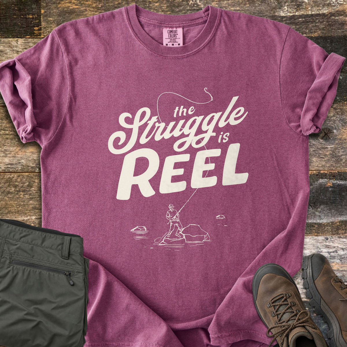 The Struggle Is Reel Comfort Colors T-shirt in Berry, laid flat. Cream text reads the Struggle is REEL under an arcing fishing line with a hook, above a sketch of an angler casting from a rock in water. Folded olive pants and brown hiking boots sit on wood planks.