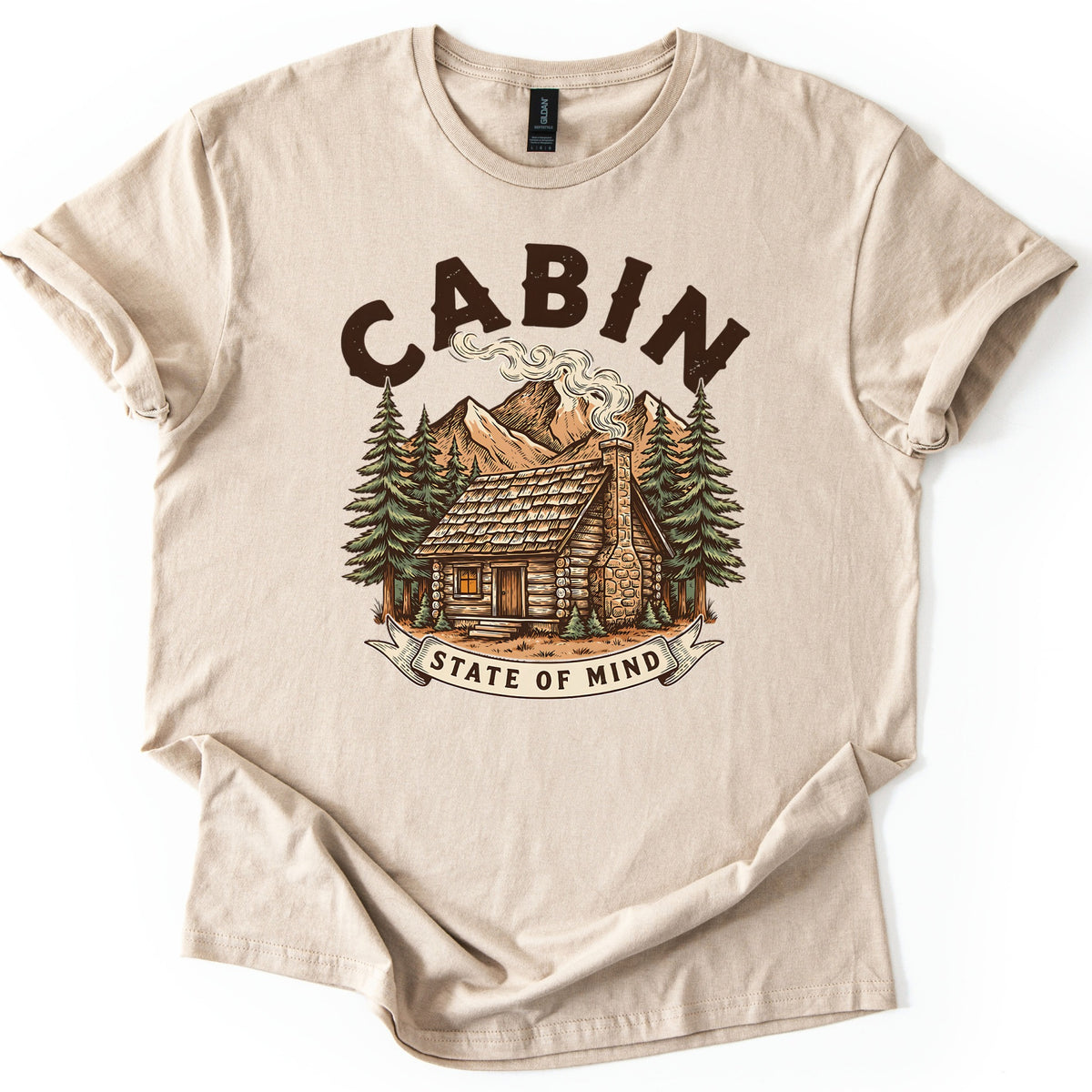 Cabin State Of Mind Comfort Colors T-shirt in Sand, laid flat. CABIN arches in distressed dark brown letters over a log cabin with a smoking stone chimney, pine trees and mountains, above a banner reading STATE OF MIND. The shirt lies on white with cuffed sleeves.