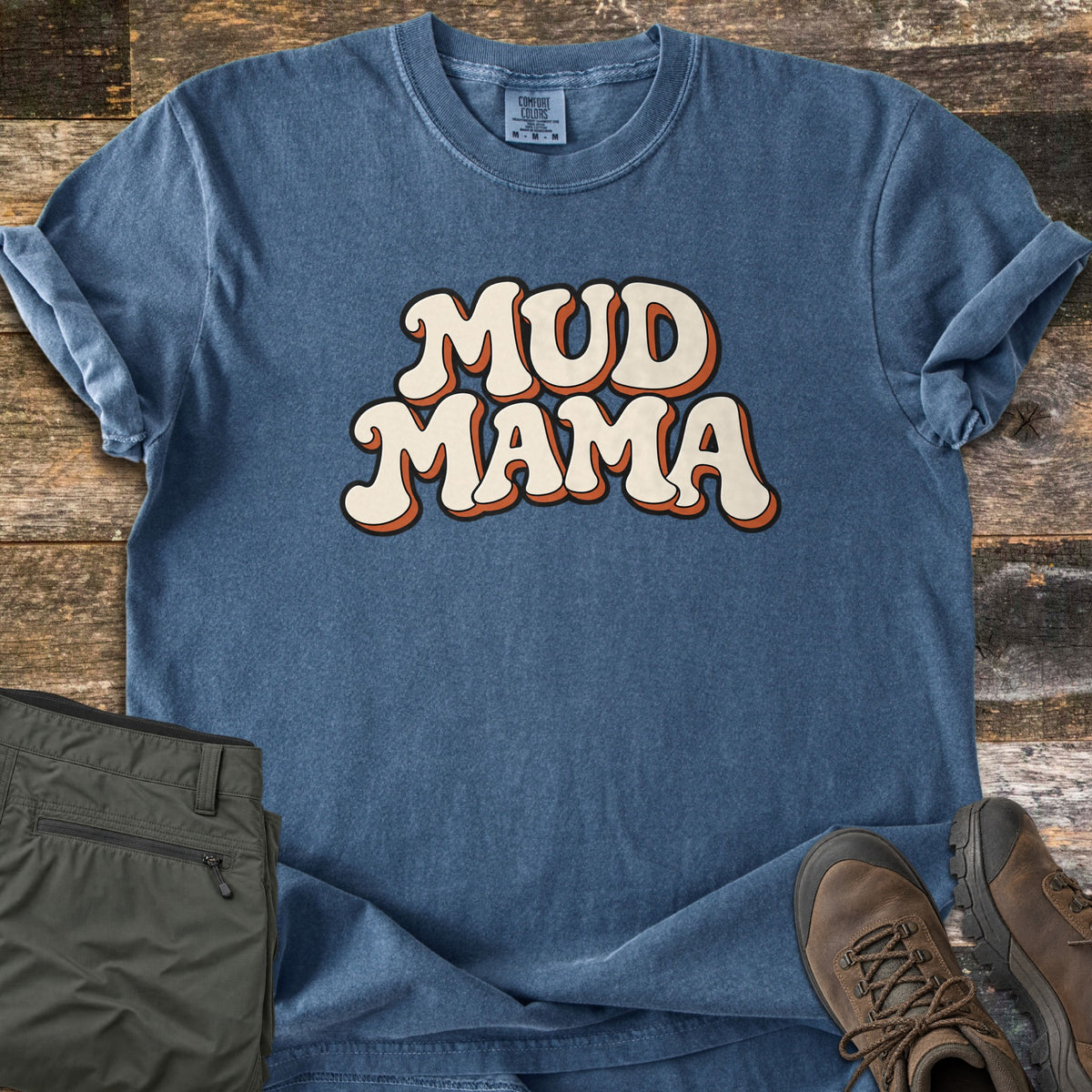 Mud Mama Comfort Colors T-shirt in Denim, laid flat. The words Mud Mama in bubbly retro cream letters with an orange drop shadow and black outline. Sleeves are rolled on a weathered wood background, with olive cargo pants and brown hiking boots beside it.