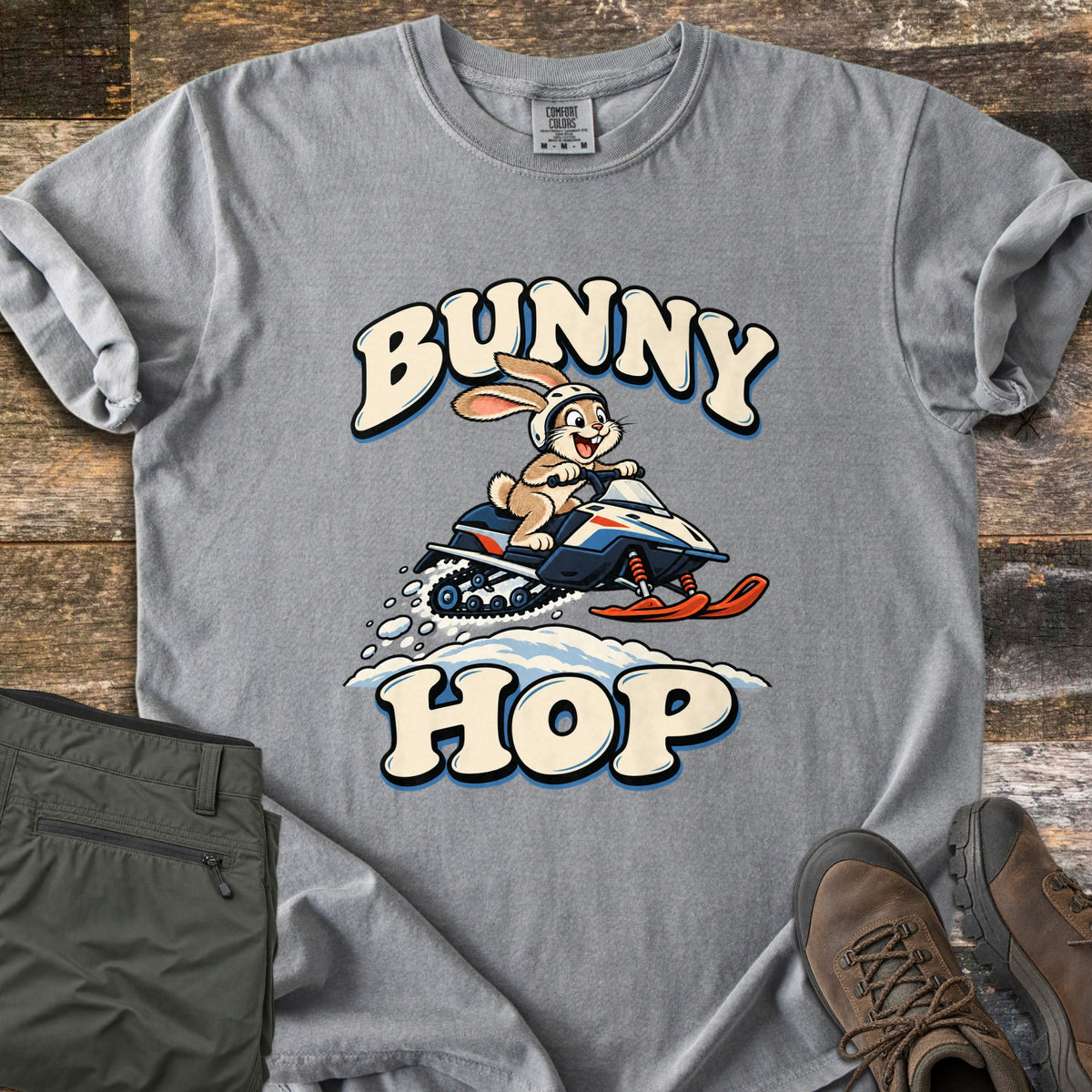 Bunny Hop Comfort Colors T-shirt in Grey, laid flat. A cartoon bunny in a white helmet rides a navy, white and orange snowmobile over a snowbank between the words Bunny Hop. Folded green pants and brown hiking boots lie beside it on a wood floor.