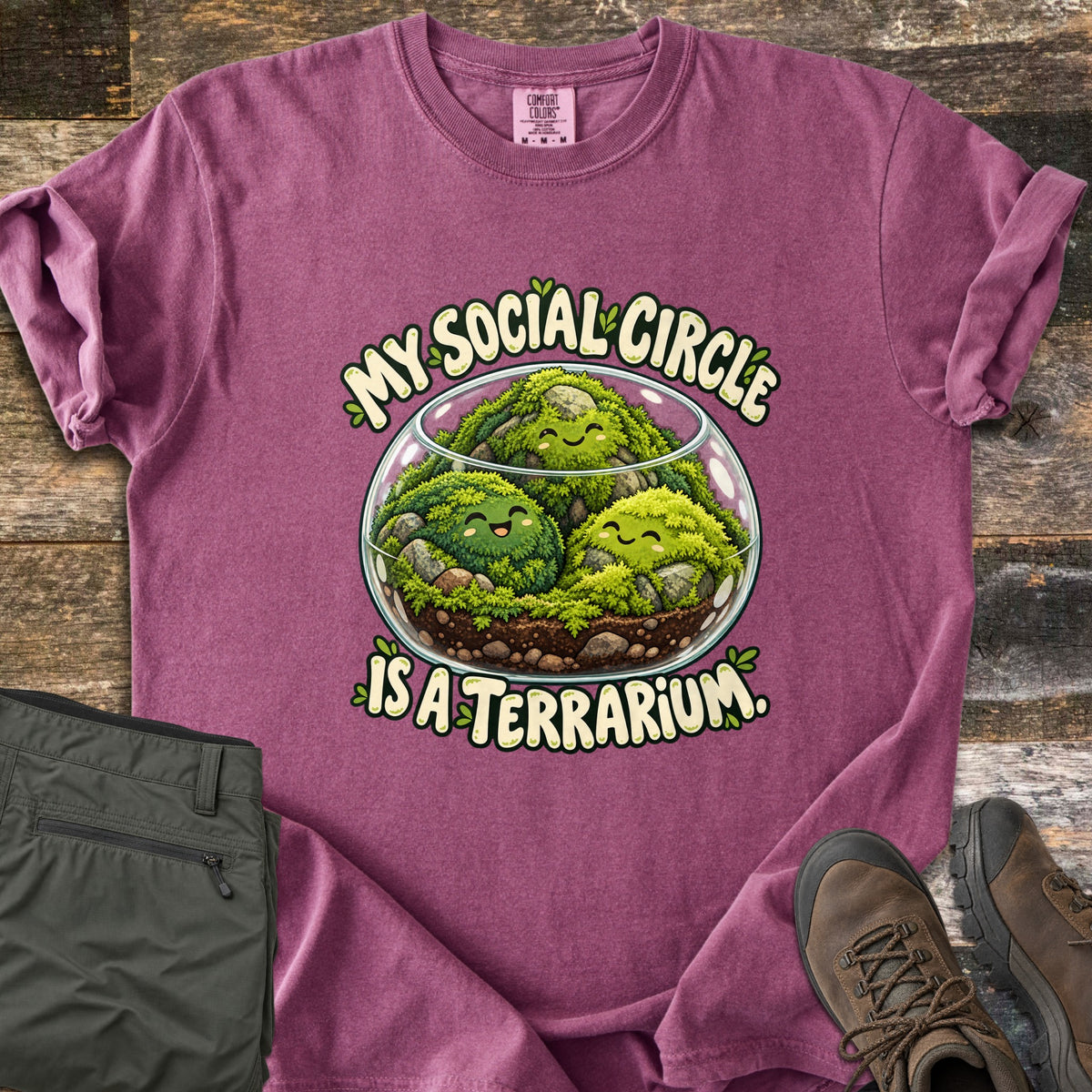 My Social Circle Is a Terrarium Comfort Colors T-shirt in Berry, laid flat. A glass bowl terrarium holds three smiling moss balls among rocks and soil, between the words My Social Circle and Is a Terrarium. Folded olive pants and brown hiking boots sit on weathered wood planks.