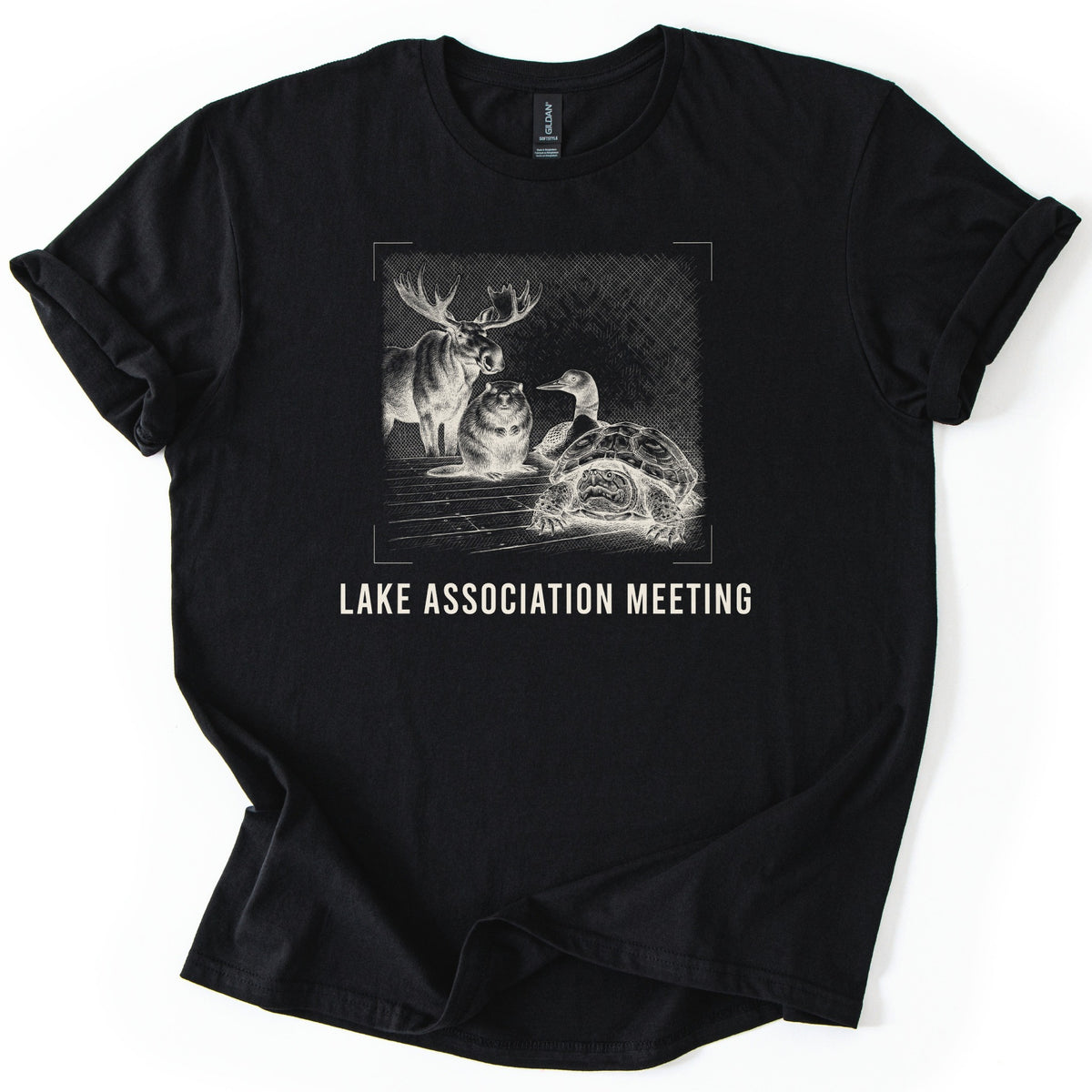 Lake Association Meeting Comfort Colors T-shirt in Black, laid flat. A cream-ink etching of a moose, a beaver, a loon, and a frowning turtle gathered on a wooden dock, framed by corner marks, above the words Lake Association Meeting. Sleeves are cuffed on a white surface.