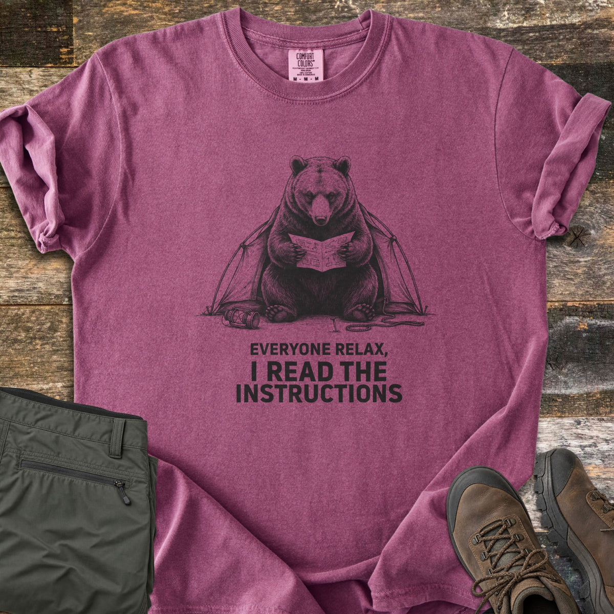Everyone Relax I Read The Instructions Comfort Colors T-shirt in Berry, laid flat. A black sketch of a bear reading instructions in front of a tent, with a tipped lantern and rope, above the words Everyone relax, I read the instructions. Olive cargo pants and brown hiking boots sit on dark wood.