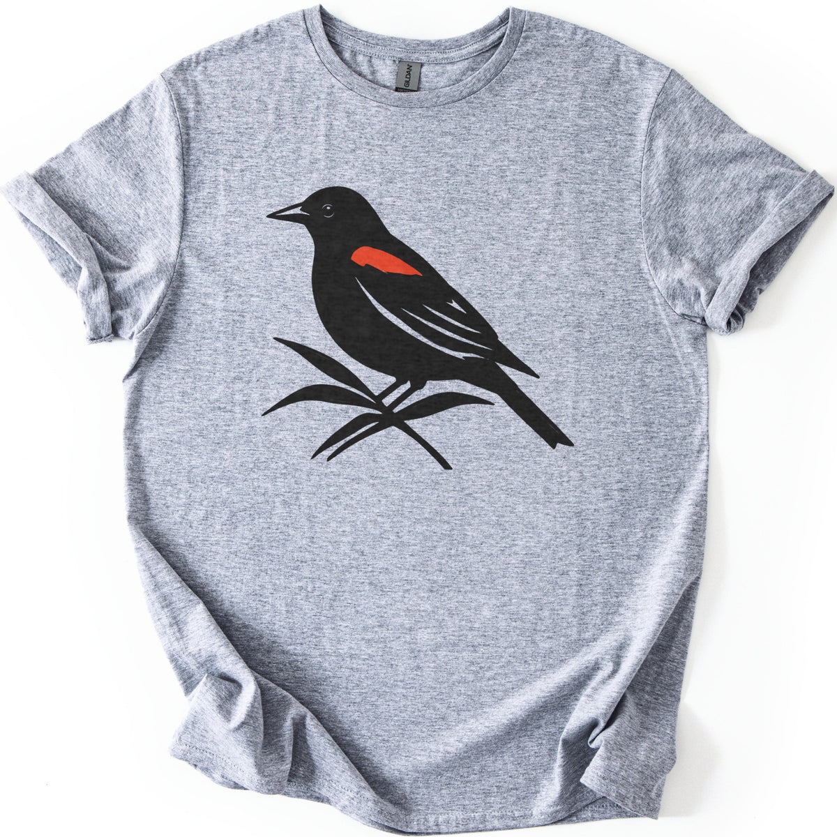 Red Winged Blackbird Comfort Colors T-shirt in Sport Grey, laid flat. A black silhouette of a red-winged blackbird with a red shoulder patch, perched on slender leaves, with no words. The shirt has rolled sleeves and lies on a white background.