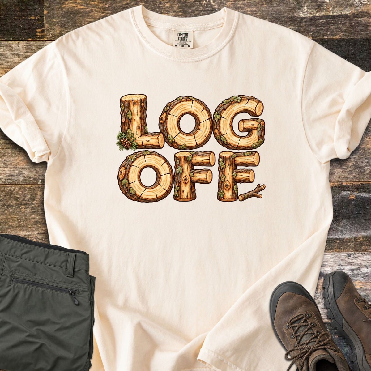 Log Off Comfort Colors T-shirt in Ivory, laid flat. The words Log Off are spelled in letters made of cut wooden logs with bark, moss and a pine cone. The shirt lies on weathered wood planks beside folded olive pants and brown hiking boots.