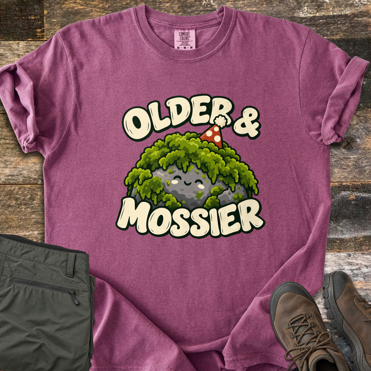 Older & Mossier Comfort Colors T-shirt in Berry, laid flat. A smiling mossy gray rock in a red polka-dot party hat is framed by the words Older & Mossier in cream letters. Sleeves are rolled, with folded olive hiking pants and brown hiking boots on weathered wood.