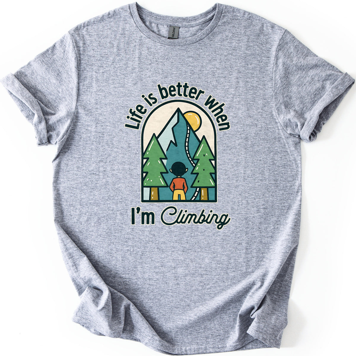Better When I'm Climbing Comfort Colors T-shirt in Sport Grey, laid flat. The words Life is better when arch over a window scene of a person facing a teal mountain with a winding trail, a yellow sun and two pine trees, with I'm Climbing below. Sleeves cuffed, on a white surface.