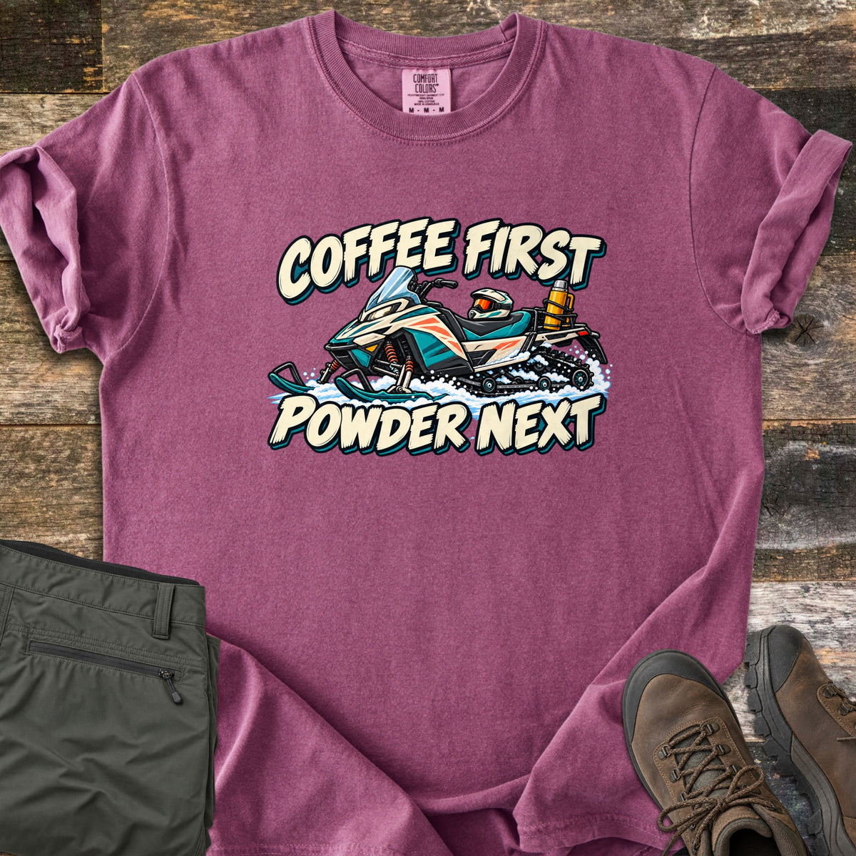 Coffee First Powder Next Comfort Colors T-shirt in Berry, laid flat. A cartoon snowmobile with a helmet on the seat and a yellow thermos strapped on back kicks up snow between the words Coffee First Powder Next. Olive hiking pants and brown boots sit on a wood floor.
