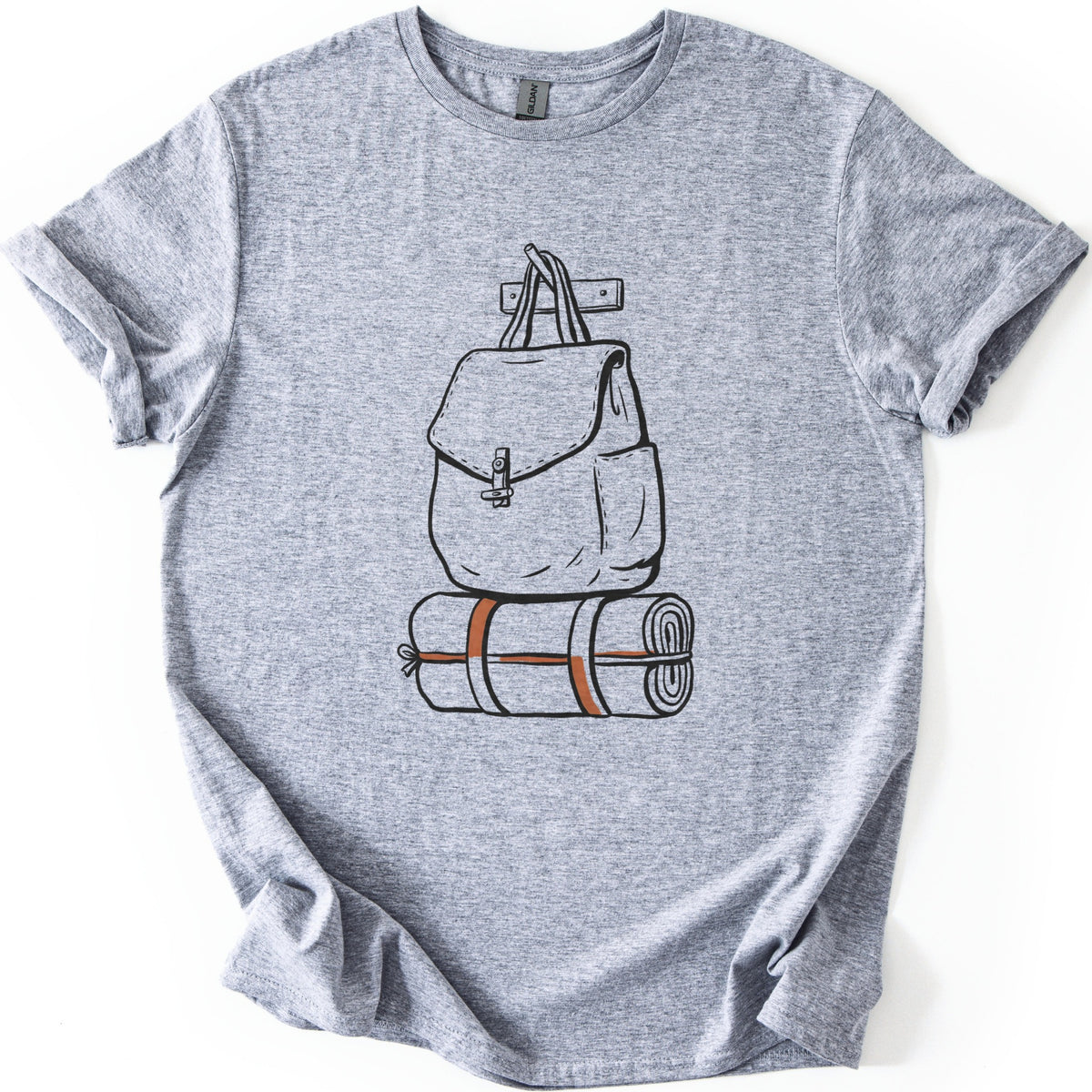 Backpack And Bedroll Comfort Colors T-shirt in Sport Grey, laid flat. A black line drawing of a canvas backpack with a toggle-clasp flap hangs by its handles from a wall peg above a rolled bedroll tied with orange straps. No words. Cuffed sleeves on a white surface.