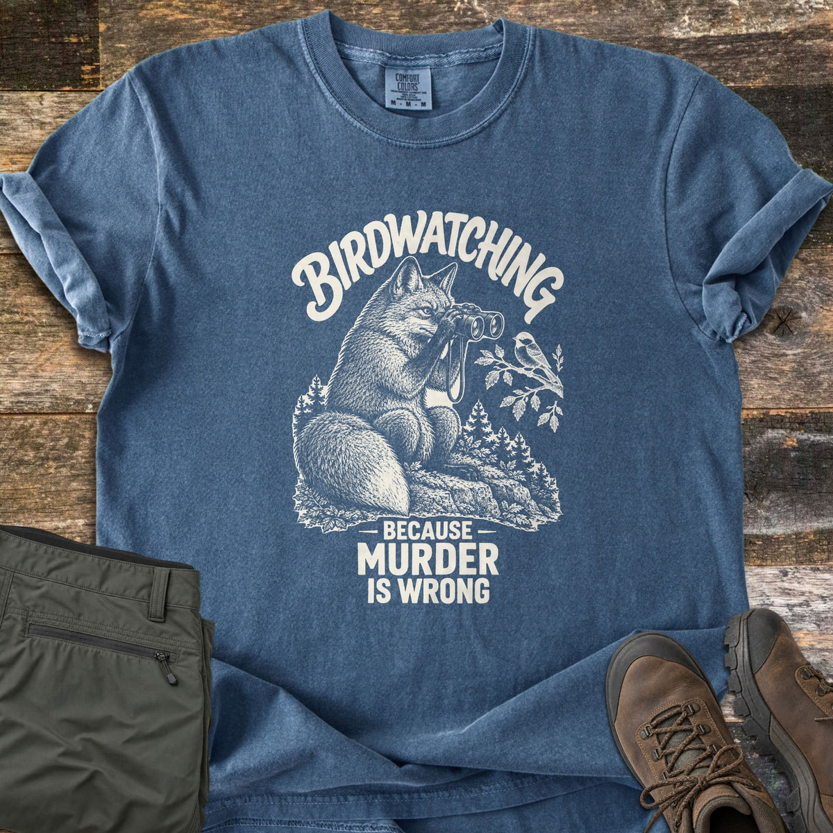 Birdwatching Because Murder Is Wrong Comfort Colors T-shirt in Denim, laid flat. A fox on a rock looks through binoculars at a small bird on a branch, with the arched word Birdwatching above and Because Murder Is Wrong below, in cream ink. Olive hiking pants and brown boots lie on weathered wood.