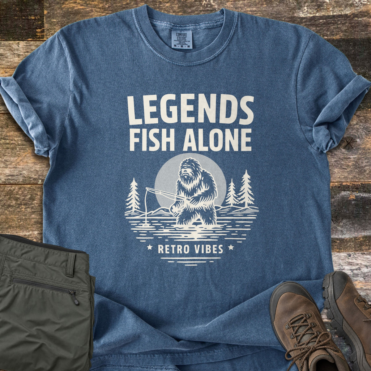 Legends Fish Alone Comfort Colors T-shirt in Denim, laid flat. Cream print of the words Legends Fish Alone above a Bigfoot fishing in a lake before a full moon and pines, with Retro Vibes between two stars below. Olive pants and brown hiking boots lie on weathered wood.