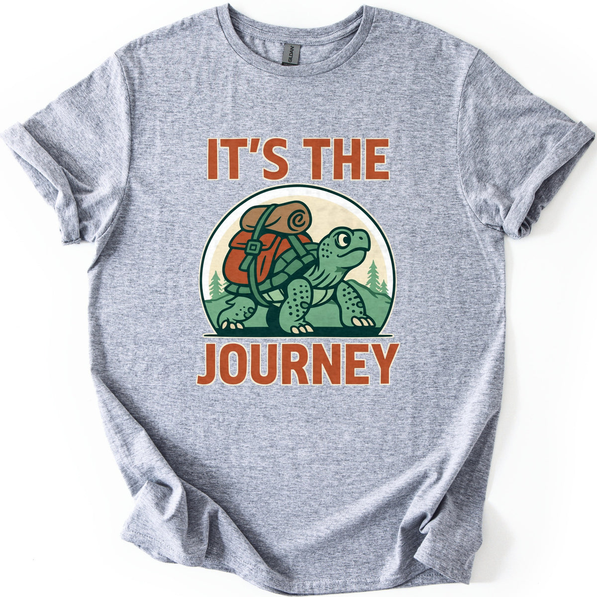 Slow Turtle Hiker Journey Comfort Colors T-shirt in Sport Grey, laid flat. A green cartoon turtle carrying a red backpack with a rolled sleeping bag walks past pine trees, framed by the words It's the Journey in rust-orange letters. Sleeves are cuffed on a white background.
