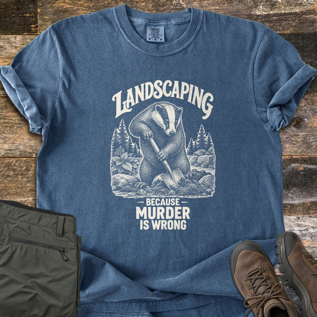 Landscaping Because Murder Is Wrong Comfort Colors T-shirt in Denim, laid flat. A cream engraving-style badger digs with a shovel among pine trees, plants and rocks under the arched word Landscaping, above Because Murder Is Wrong. Set on weathered wood beside olive hiking pants and brown boots.