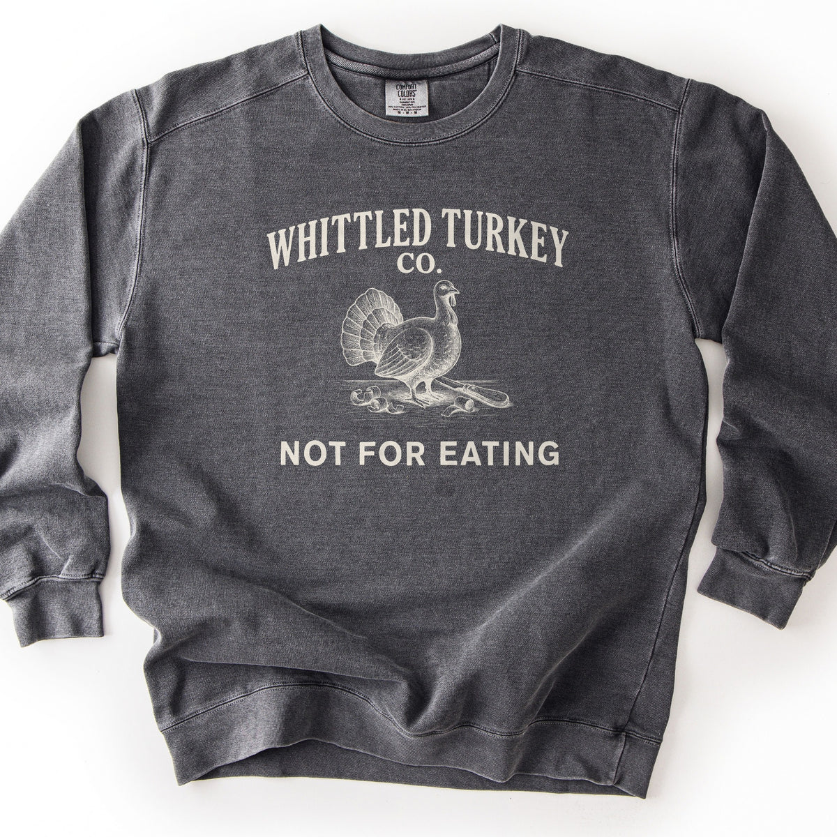 Whittled Turkey Co Comfort Colors crewneck sweatshirt in Pepper, laid flat. Cream print of the words Whittled Turkey Co. arched over an engraved-style turkey standing beside a carving knife and wood shavings, with Not For Eating below. The sweatshirt lies on a white background.