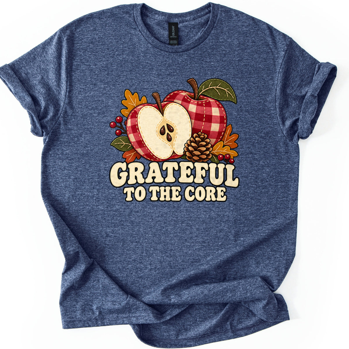 Grateful to the Core Comfort Colors T-shirt in Heather Navy, laid flat. Red and cream plaid apples, one cut open to show seeds, are surrounded by a pinecone, autumn leaves and red berries above the words Grateful to the Core. Shown with rolled sleeves on a white background.