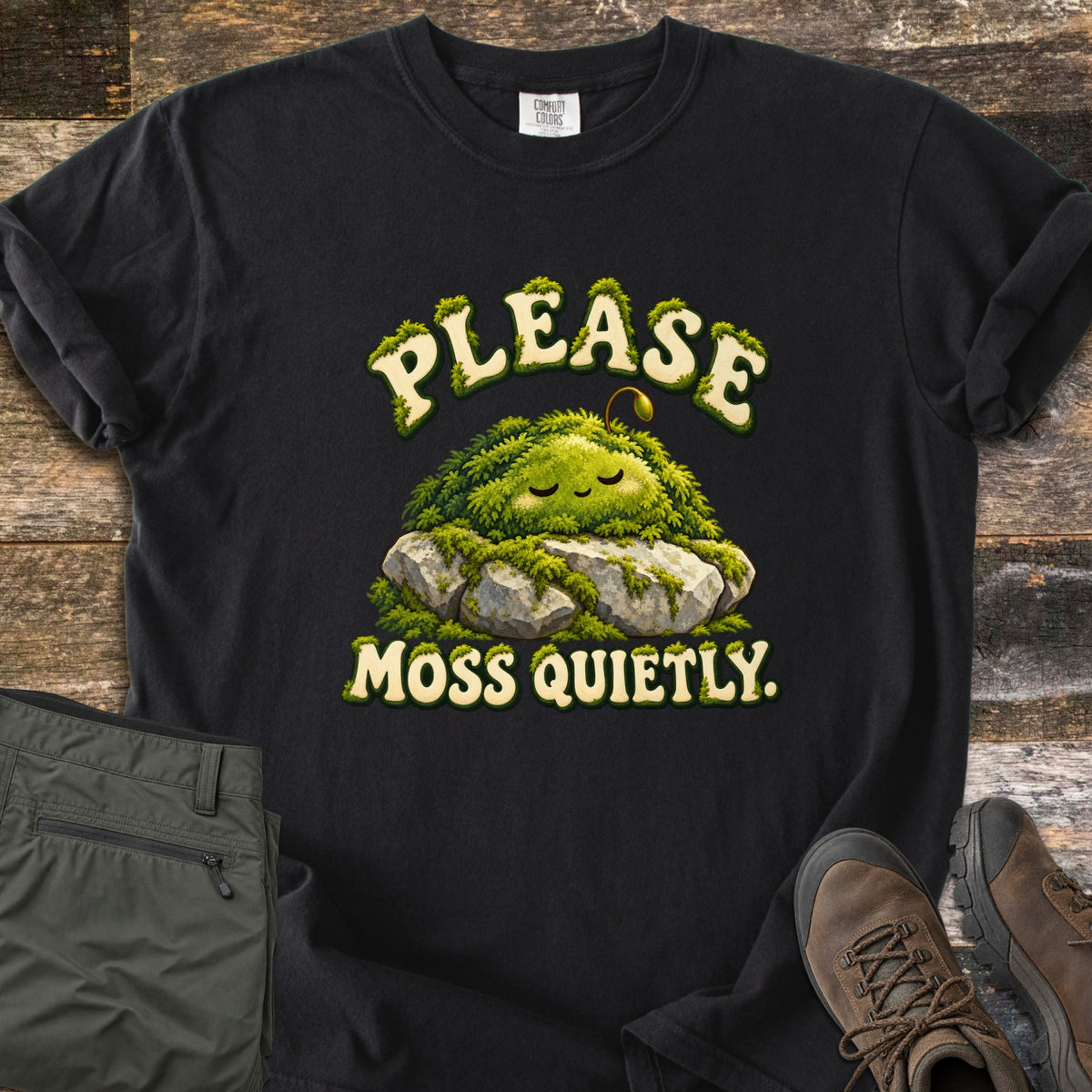 Please Moss Quietly Comfort Colors T-shirt in Black, laid flat. A sleepy, smiling clump of moss with a tiny sprout rests on gray rocks, with PLEASE arched above and MOSS QUIETLY. below in moss-covered letters. Olive hiking pants and brown hiking boots lie on weathered wood.