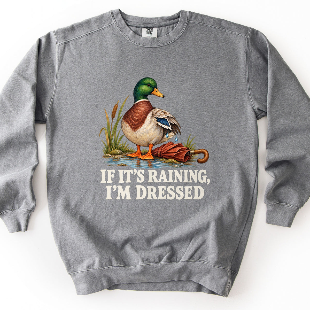 If It's Raining, I'm Dressed Comfort Colors crewneck sweatshirt in Grey, laid flat. A mallard duck with water dripping from its wing stands in a puddle by cattails beside a closed red umbrella, above the words If It's Raining, I'm Dressed in cream. Shown on a white background.