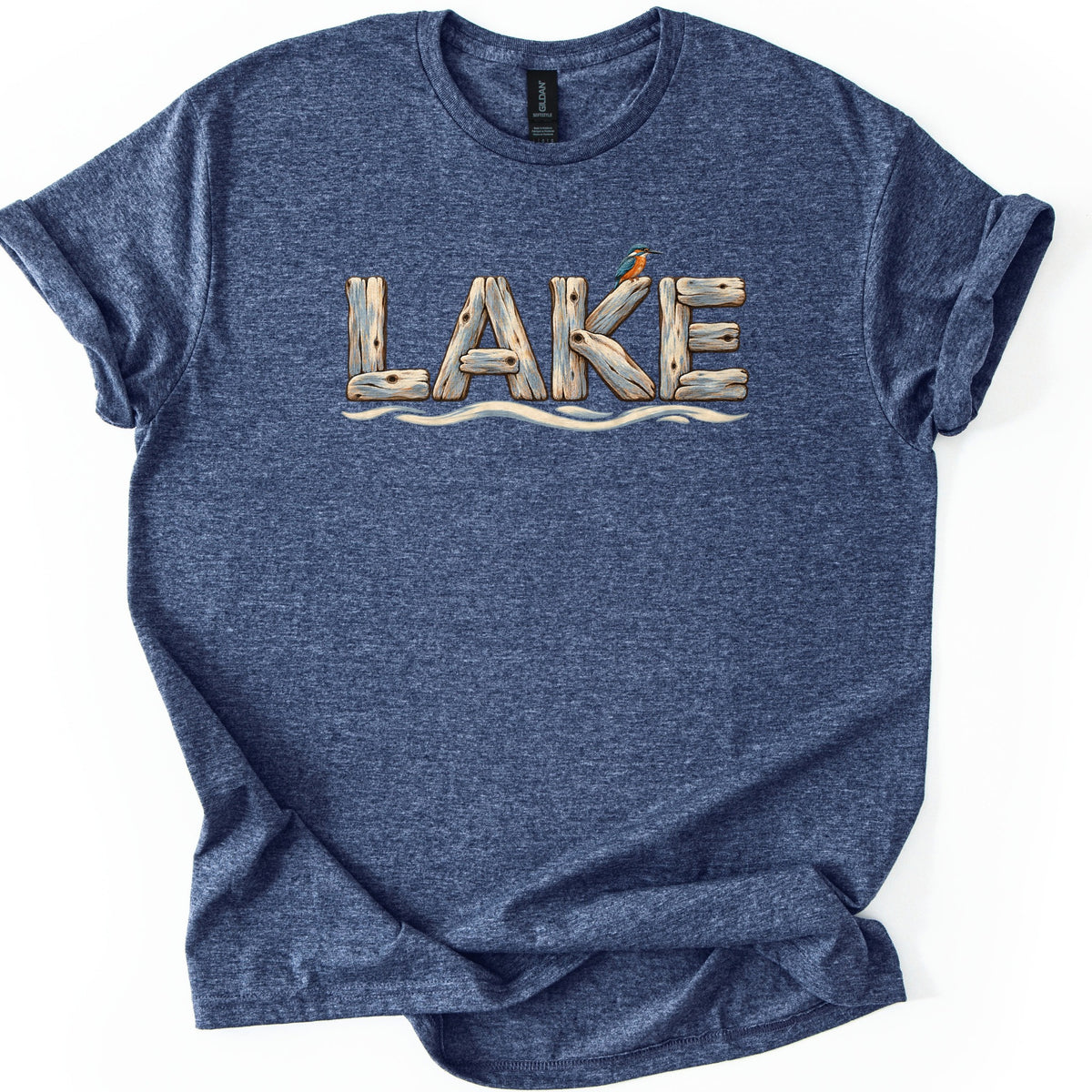 Lake Comfort Colors T-shirt in Heather Navy, laid flat. The word LAKE is spelled in weathered driftwood planks, with a small blue and orange kingfisher perched on the K and wavy water lines below. The shirt lies on a white surface with sleeves cuffed.