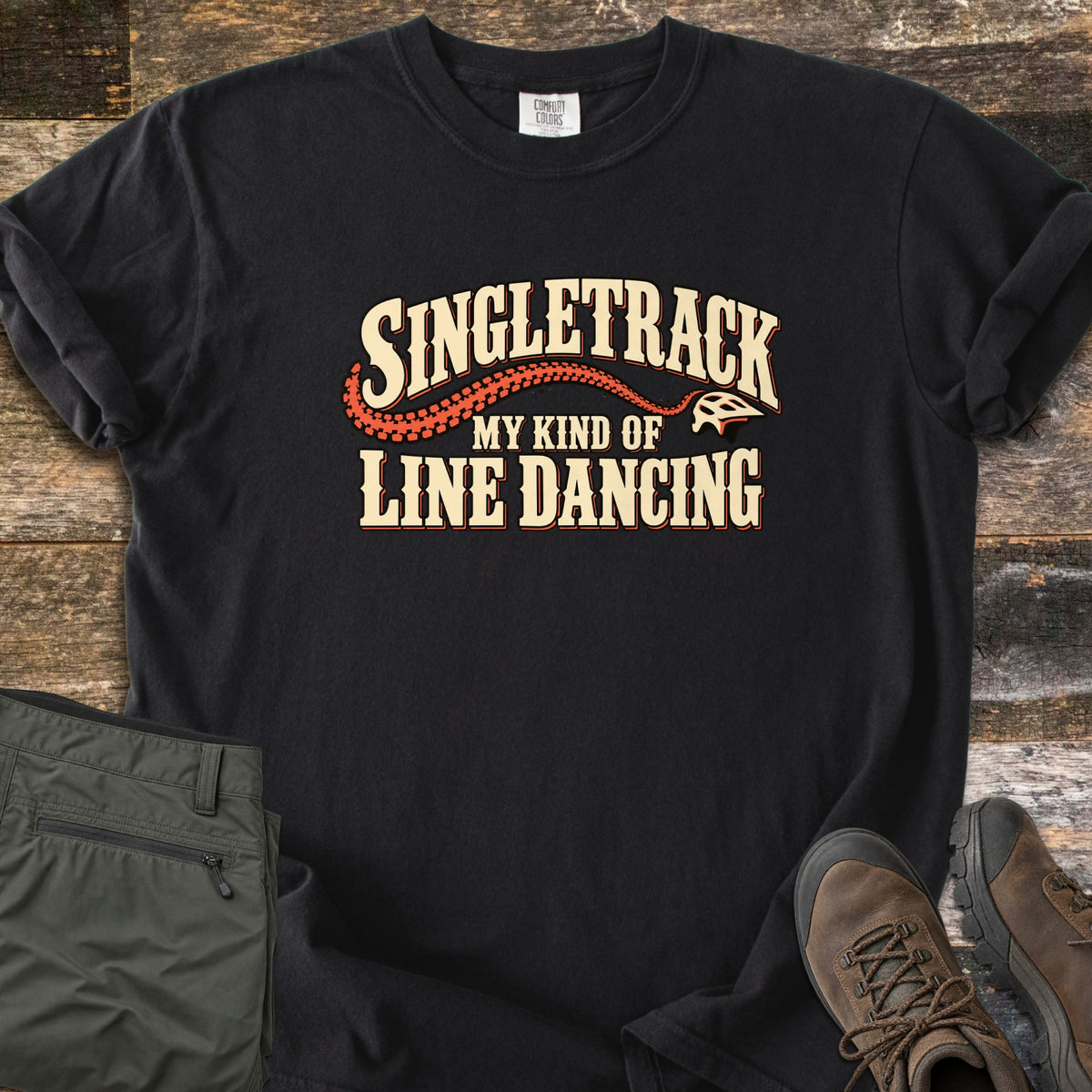 Singletrack My Kind of Line Dancing Comfort Colors T-shirt in Black, laid flat. Cream western-style lettering reads Singletrack, My Kind of Line Dancing, with an orange tire track curving to a bike helmet. Shown on weathered wood with folded green hiking pants and brown hiking boots.