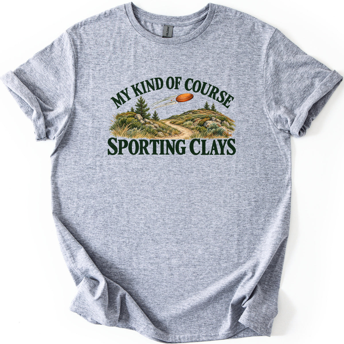 My Kind of Course Comfort Colors T-shirt in Sport Grey, laid flat. Dark green text My Kind of Course arcs over an orange clay target flying above hills with pines, rocks and a dirt trail, with Sporting Clays below. The shirt lies on a white background with sleeves rolled.