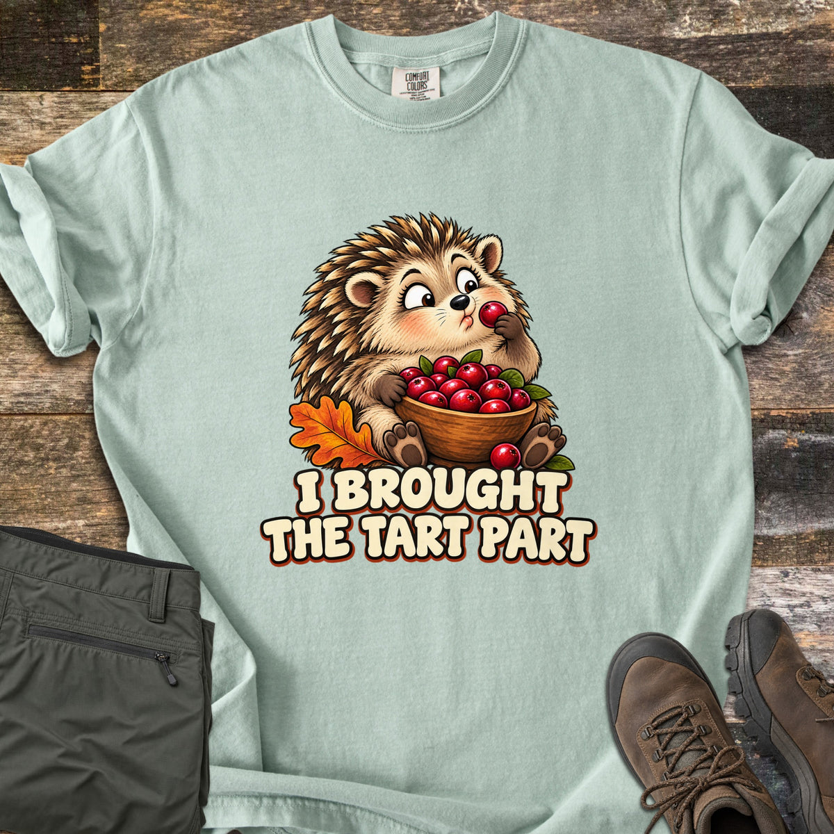I Brought the Tart Part Comfort Colors T-shirt in Bay, laid flat. A cartoon hedgehog nibbles a cranberry while holding a wooden bowl of cranberries beside an orange oak leaf, above the words I Brought the Tart Part. Olive pants and brown hiking boots sit on a wood background.