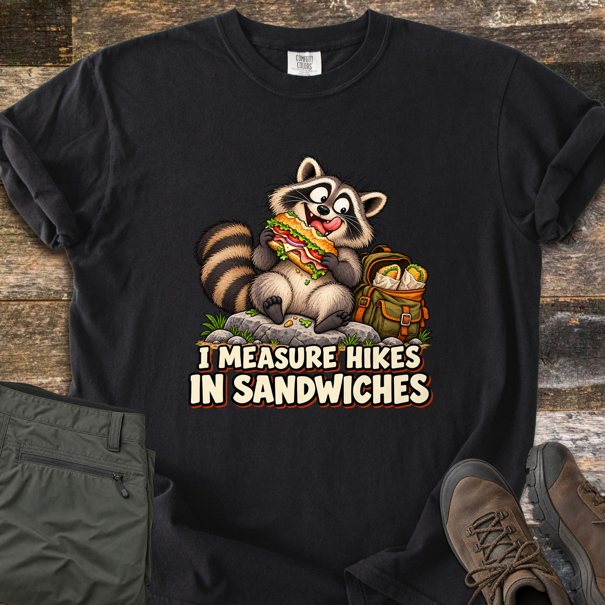 I Measure Hikes In Sandwiches Comfort Colors T-shirt in Black, laid flat. A cartoon raccoon sits on a rock biting a big sub sandwich beside a green backpack holding wrapped burritos, above the words I Measure Hikes In Sandwiches. Hiking pants and boots lie on a wood floor beside it.