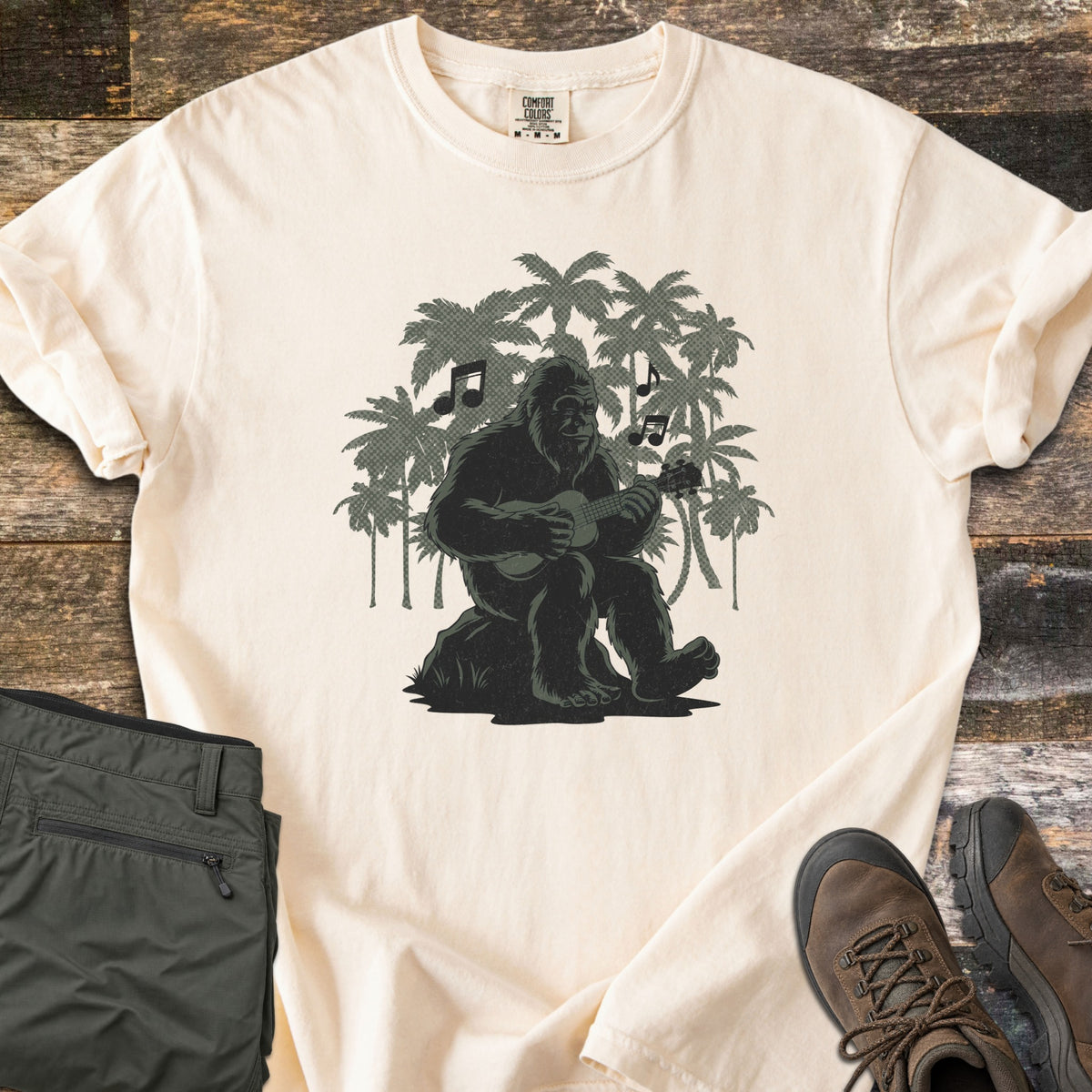 Bigfoot Ukulele Comfort Colors T-shirt in Ivory, laid flat. A Bigfoot sits on a rock playing a ukulele, with music notes and green palm tree silhouettes behind him. Olive hiking pants and brown hiking boots lie beside it on weathered wood planks.