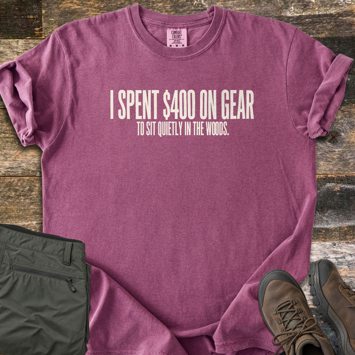 Gear To Sit Quietly Comfort Colors T-shirt in Berry, laid flat. Cream text reads I SPENT $400 ON GEAR, with TO SIT QUIETLY IN THE WOODS. below. Sleeves are rolled on a rustic wood surface, beside folded olive hiking pants and brown hiking boots.