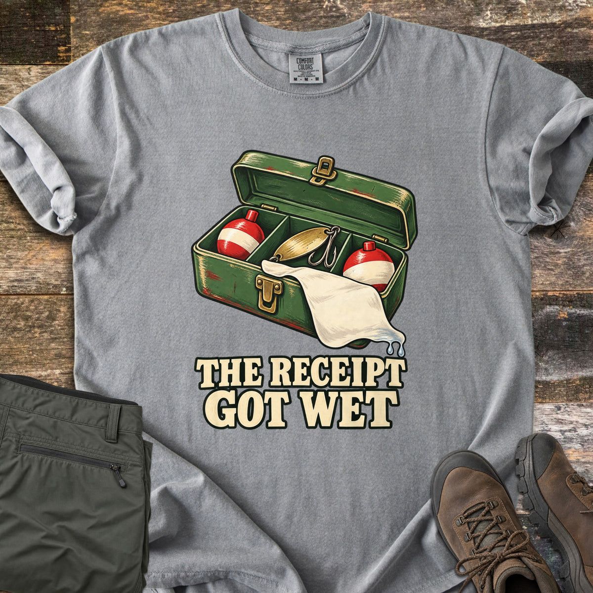 The Receipt Got Wet Comfort Colors T-shirt in Grey, laid flat. An open green tackle box holds red and white bobbers and a gold spoon lure, with a dripping white paper hanging out, above the words The Receipt Got Wet. Olive pants and brown hiking boots sit on weathered wood.
