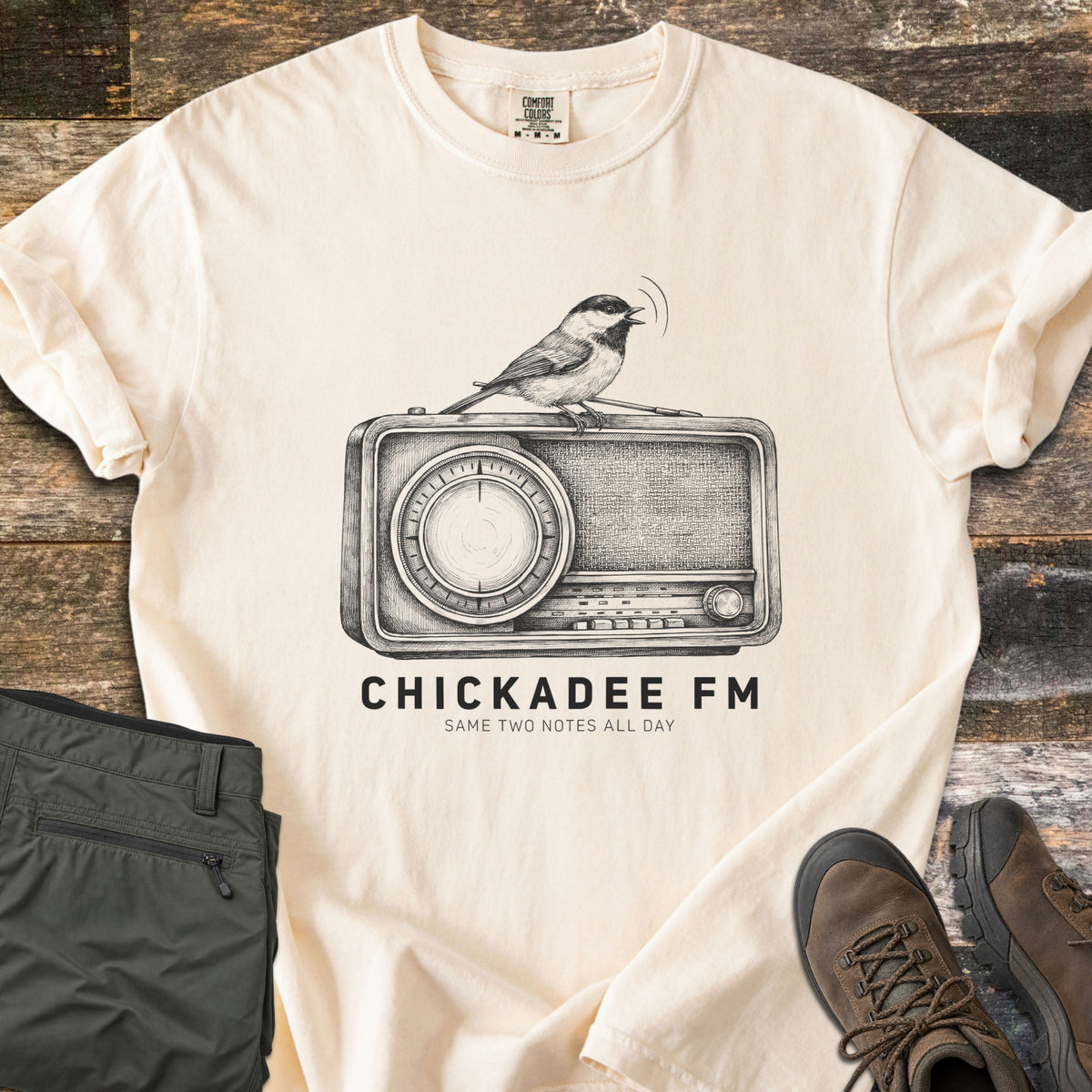 Chickadee FM Comfort Colors T-shirt in Ivory, laid flat. A sketched chickadee singing atop a vintage radio, above the words CHICKADEE FM and Same Two Notes All Day in black. Sleeves rolled, on weathered wood with folded green hiking pants and brown hiking boots.