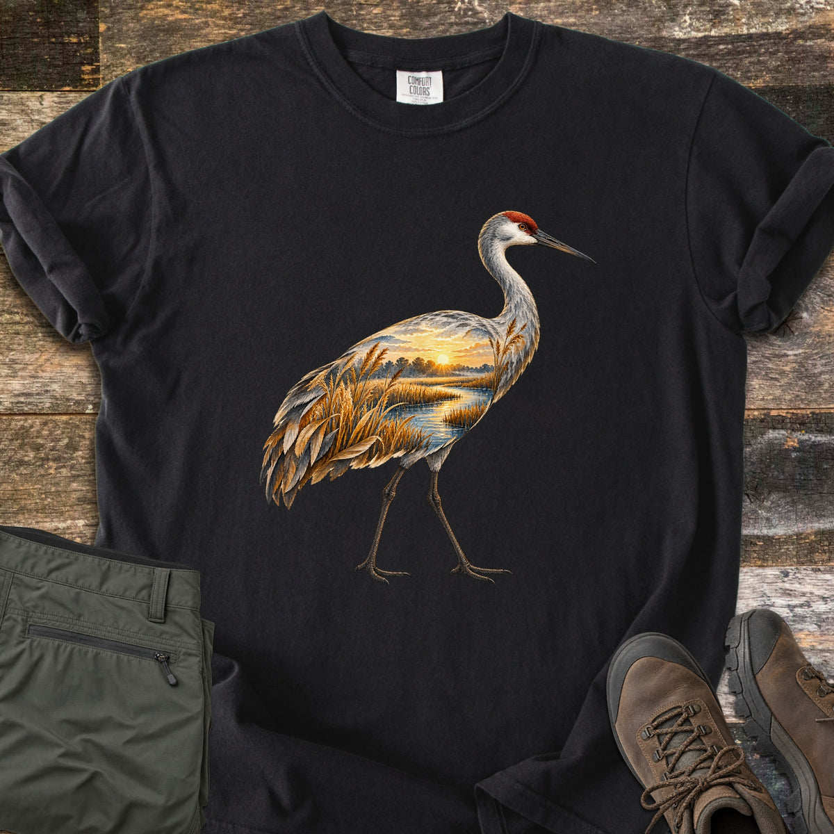 Crane Marsh Sunrise Comfort Colors T-shirt in Black, laid flat. A sandhill crane with a red crown walks across the chest, its body filled with a sunrise marsh scene of golden reeds and a winding stream. Rolled sleeves; gray pants and brown hiking boots sit on weathered wood.
