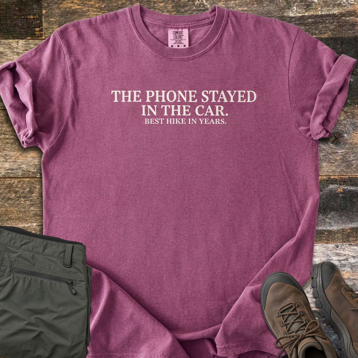 Phone Stayed In The Car Comfort Colors T-shirt in Berry, laid flat. Cream serif text reads THE PHONE STAYED IN THE CAR. with BEST HIKE IN YEARS. in smaller letters below. Sleeves are rolled on a wood plank surface beside olive hiking pants and brown hiking boots.