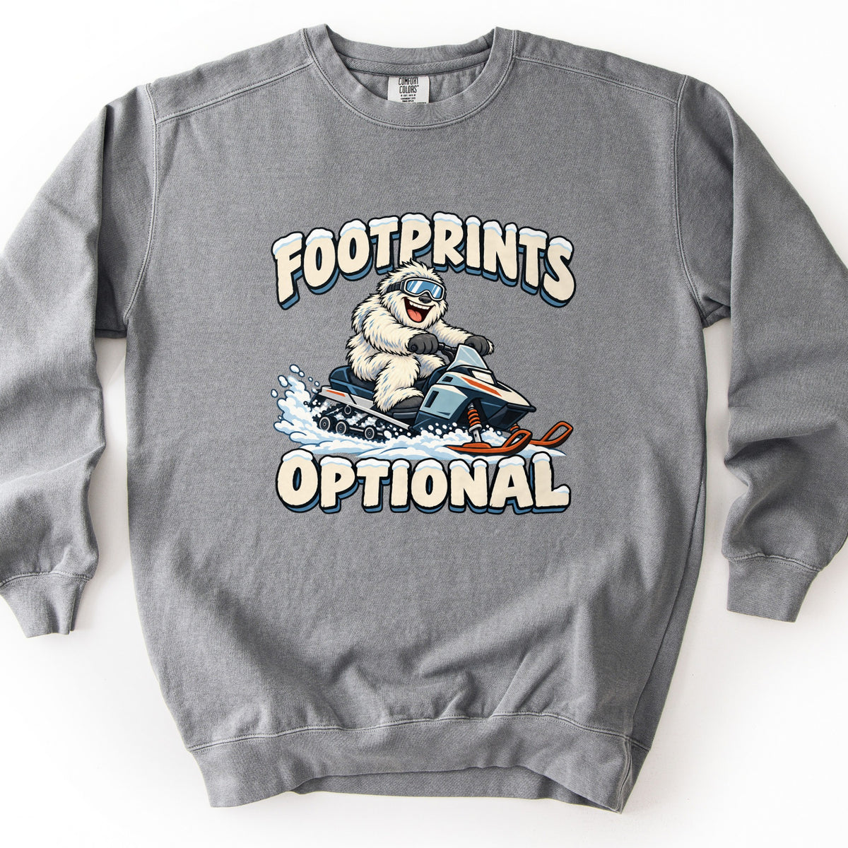Footprints Optional Comfort Colors crewneck sweatshirt in Grey, laid flat. A cartoon white yeti in blue ski goggles rides a gray and orange snowmobile through powder, with snow-capped cream letters reading FOOTPRINTS above and OPTIONAL below. Shown on a white background.