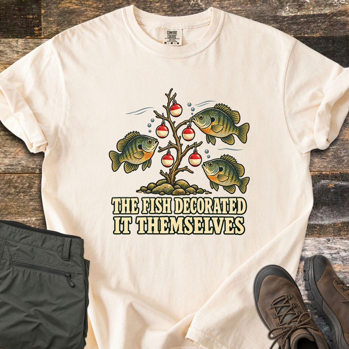 The Fish Decorated It Themselves Comfort Colors T-shirt in Ivory, laid flat. Three bluegill fish hang red and white fishing bobbers like ornaments on a bare underwater branch above the words The Fish Decorated It Themselves. Olive pants and brown hiking boots sit on weathered wood.