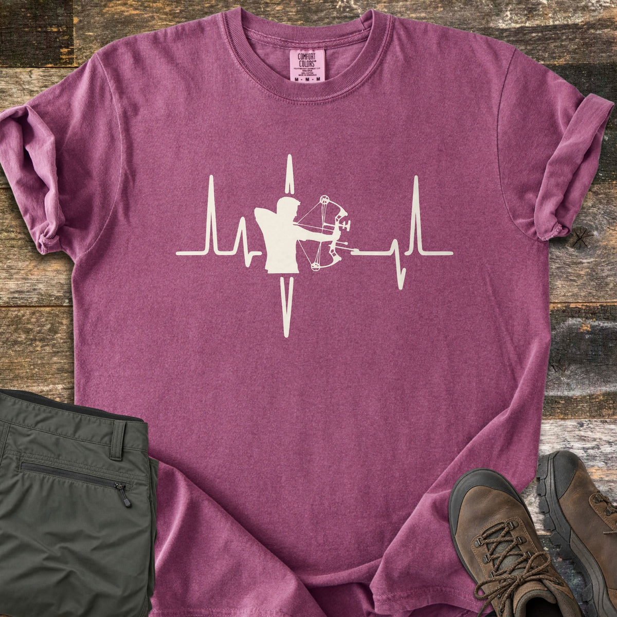 Compound Bow Heartbeat Comfort Colors T-shirt in Berry, laid flat. A cream heartbeat line with the silhouette of an archer drawing a compound bow at its center. Sleeves are rolled on a wood plank floor, with olive cargo pants and brown hiking boots beside it.