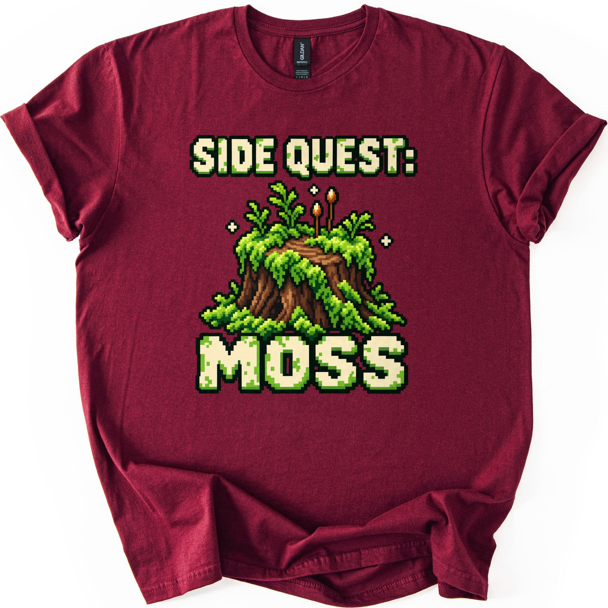 Side Quest Moss Comfort Colors T-shirt in Maroon, laid flat. The words Side Quest: in cream pixel letters sit above a moss-covered tree stump with ferns and two spore stalks, and Moss is printed below. The shirt lies on a white background with sleeves rolled.
