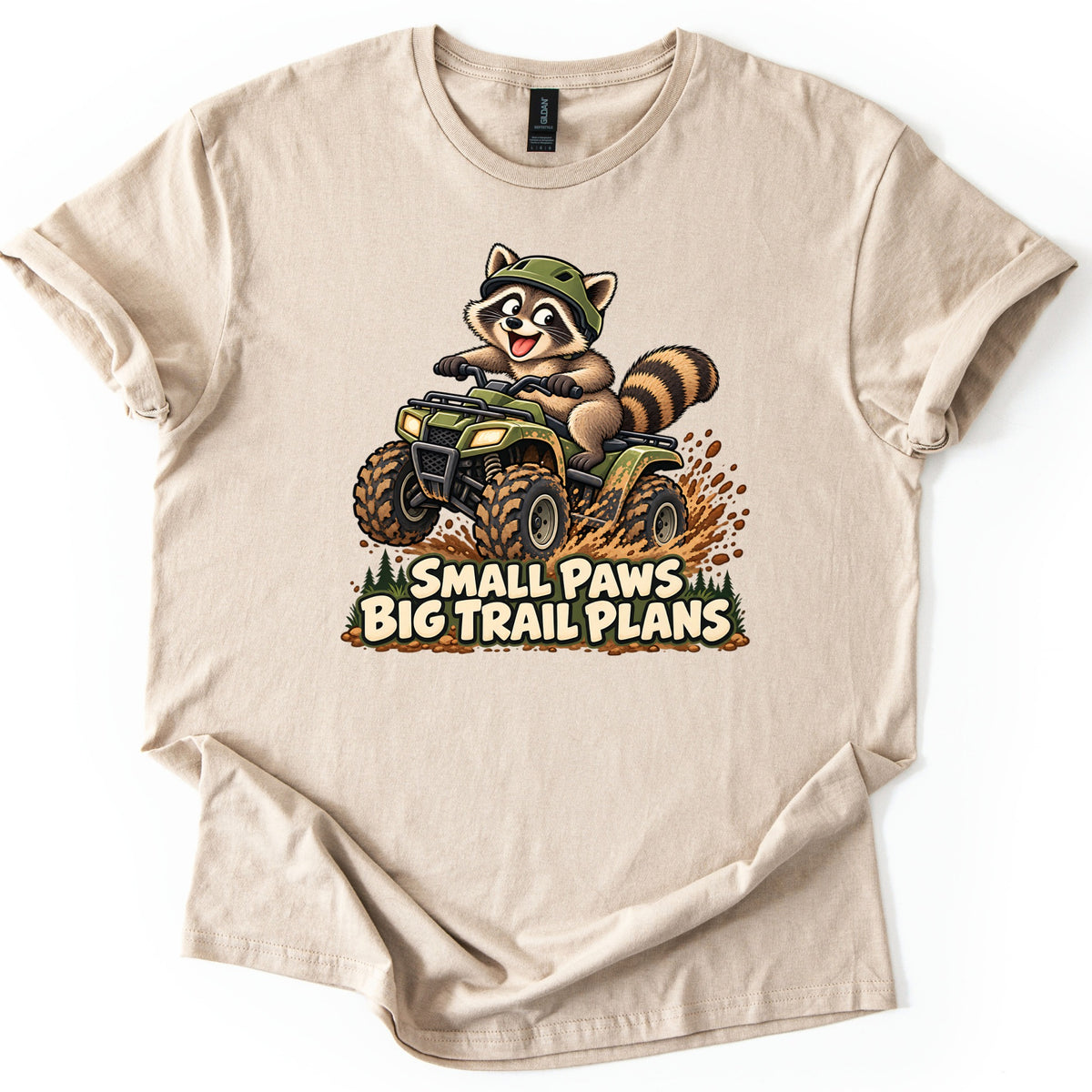 Small Paws Big Trail Plans Comfort Colors T-shirt in Sand, laid flat. A cartoon raccoon in a green helmet rides a muddy green ATV, kicking up dirt beside pine trees, above the words Small Paws Big Trail Plans. The shirt lies on a white background with cuffed sleeves.