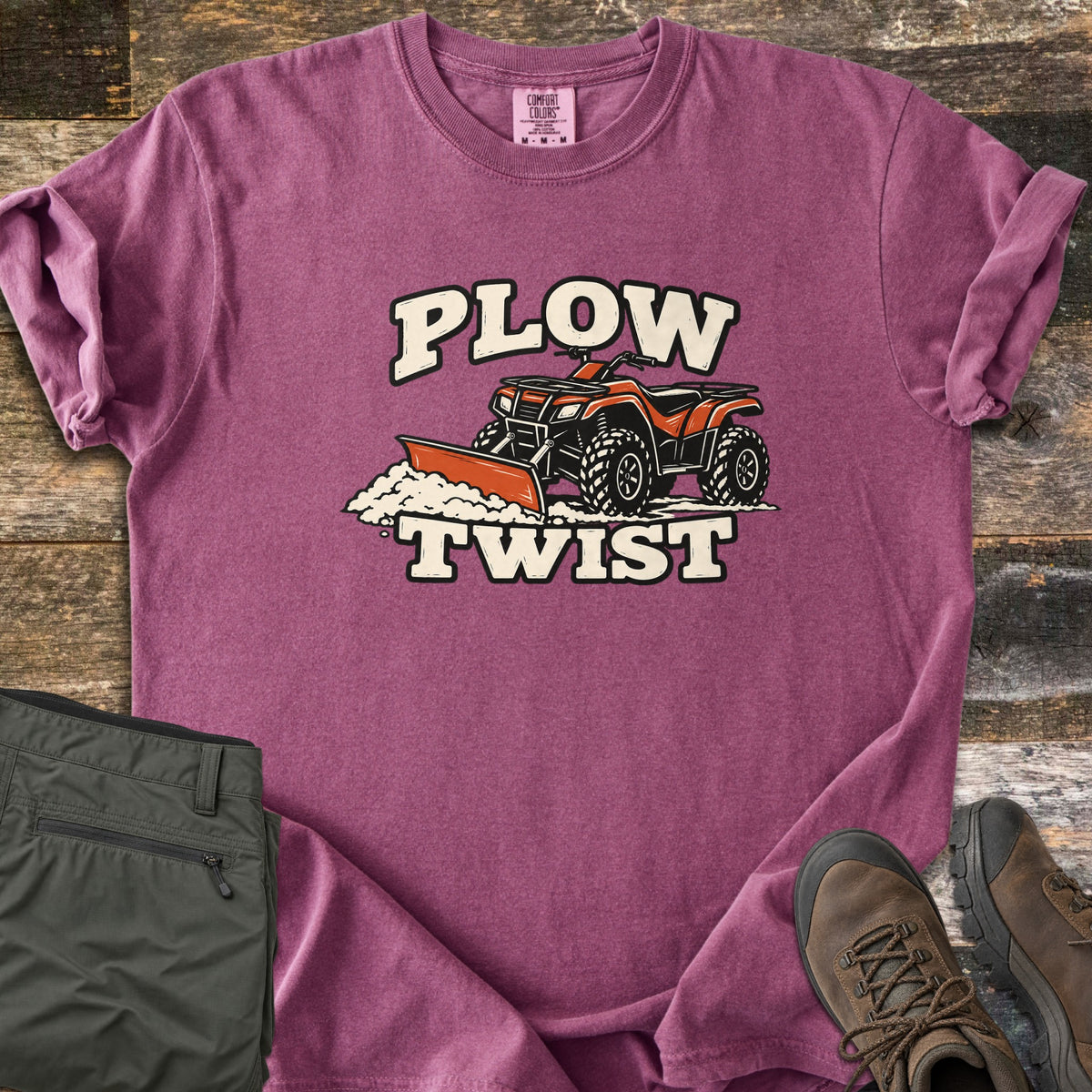 Plow Twist Comfort Colors T-shirt in Berry, laid flat. An orange ATV with a snow plow blade pushes a pile of snow between the words Plow Twist in cream lettering. Folded olive cargo pants and brown hiking boots sit beside it on weathered wood planks.
